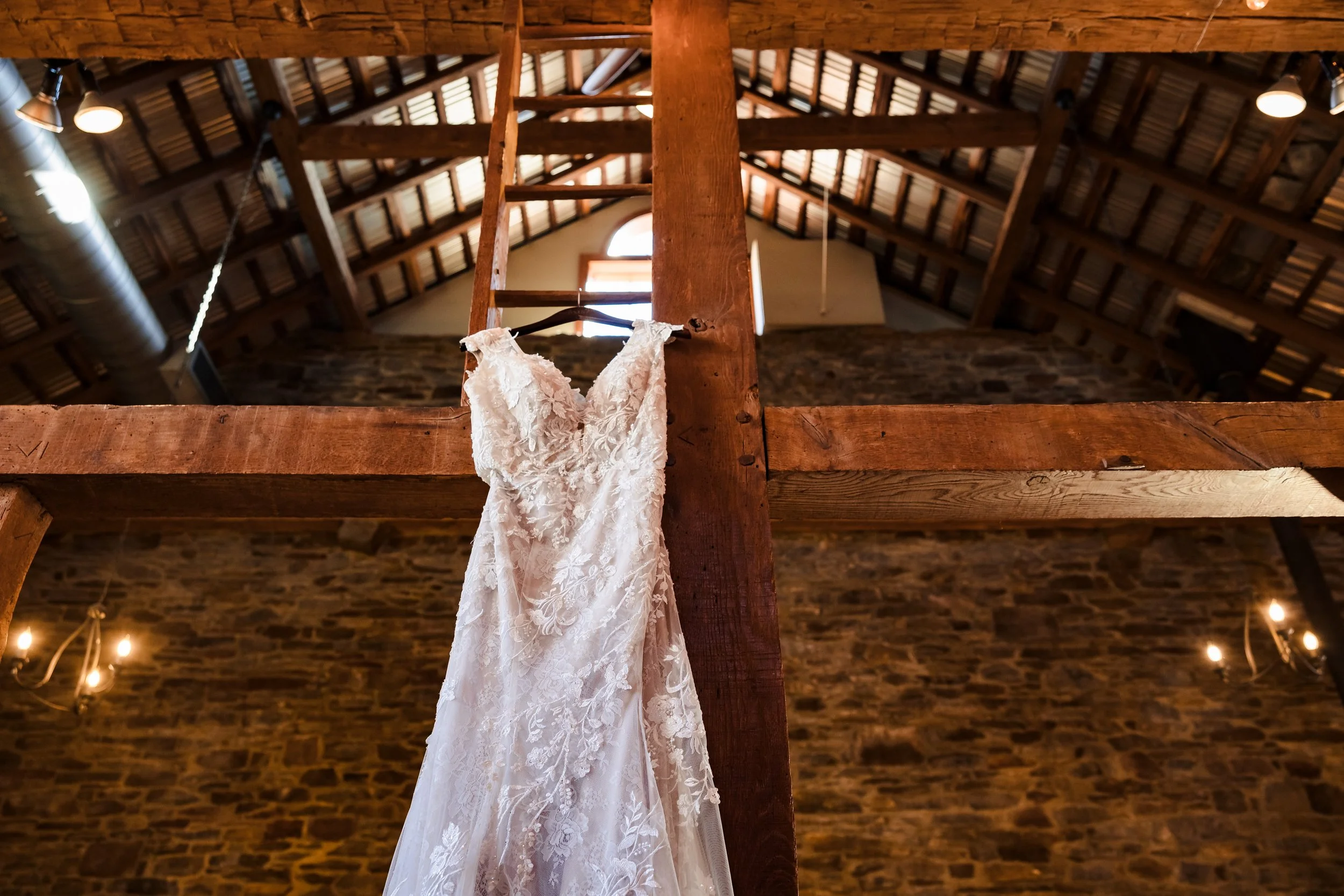 A white wedding dress with lace details hanging on a black hanger from a wooden beam inside a rustic barn with exposed brick walls, wooden ceiling beams, and warm lighting.