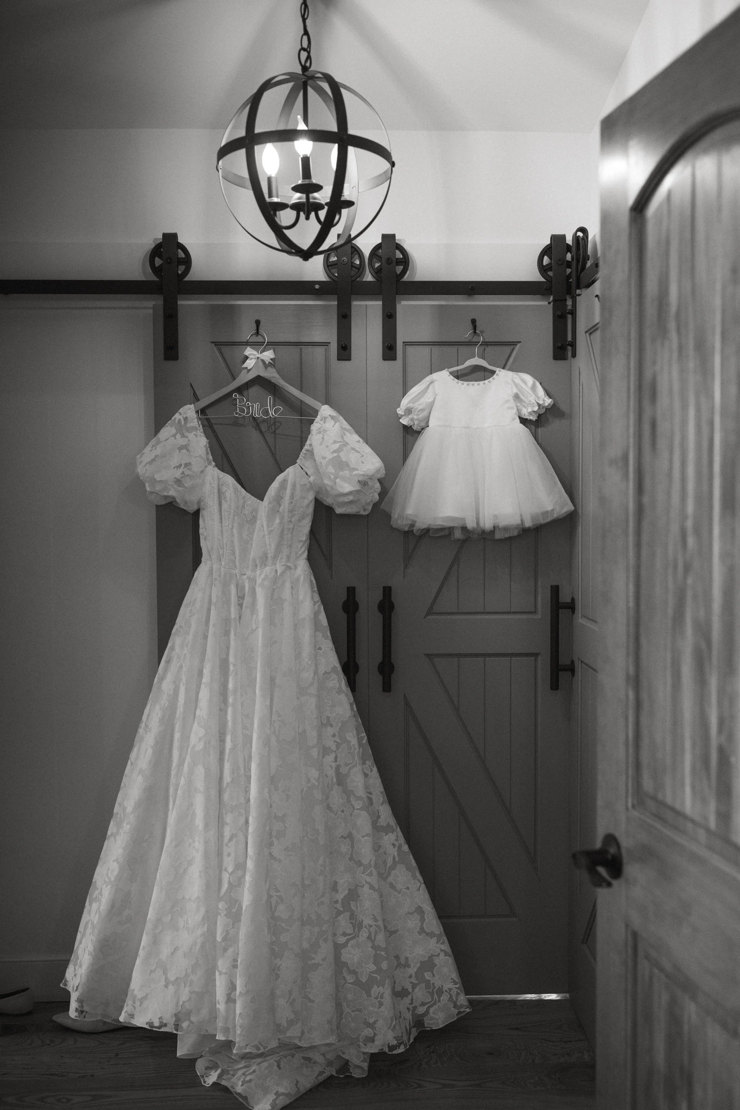 A black and white photo of a bridal dress hanging on a hanger with a sign that says 'Bride' on a wooden door. There is a chandelier hanging from the ceiling above the dress.