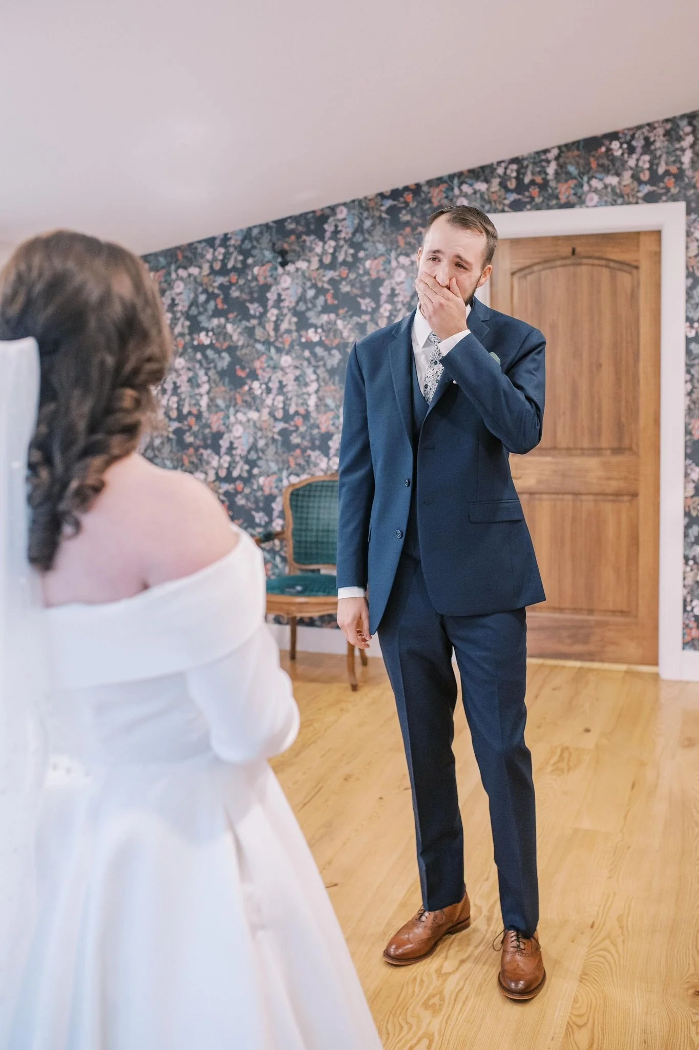 A man in a blue suit covering his mouth with his hand, appearing emotional, standing in front of a woman in a white dress. The woman is seated, with her back to the camera, in a room with floral wallpaper and wooden flooring.
