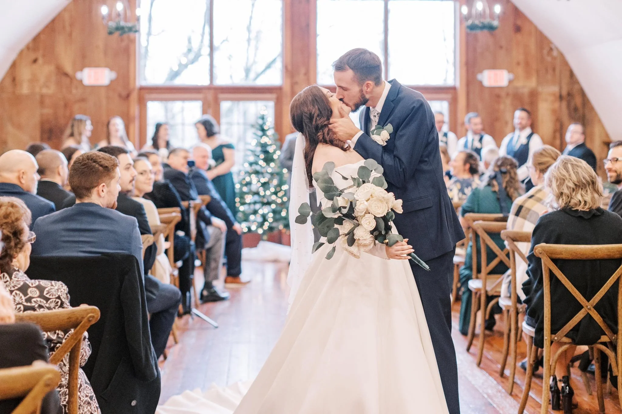 A bride and groom kiss during their wedding ceremony in a wood-paneled indoor venue, with seated guests watching and a decorated Christmas tree in the background.