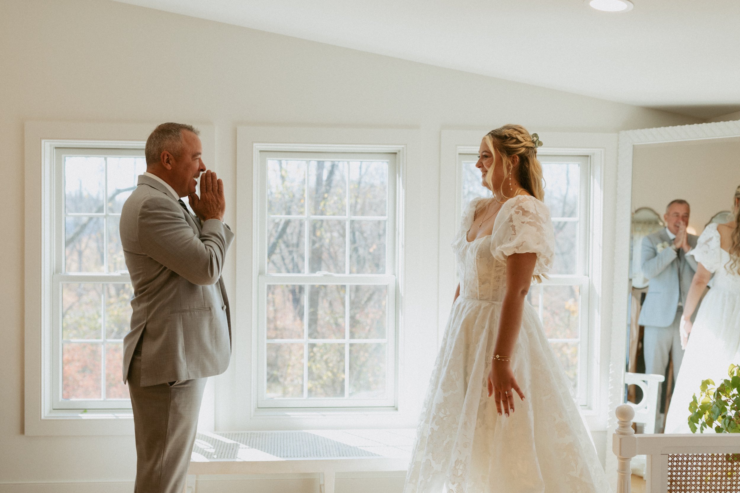 A man in a light gray suit with a white shirt and tie is smiling and covering his mouth with his hands, looking at a woman in a white wedding dress with lace details. They are inside a bright room with large windows showing trees outside. In the back