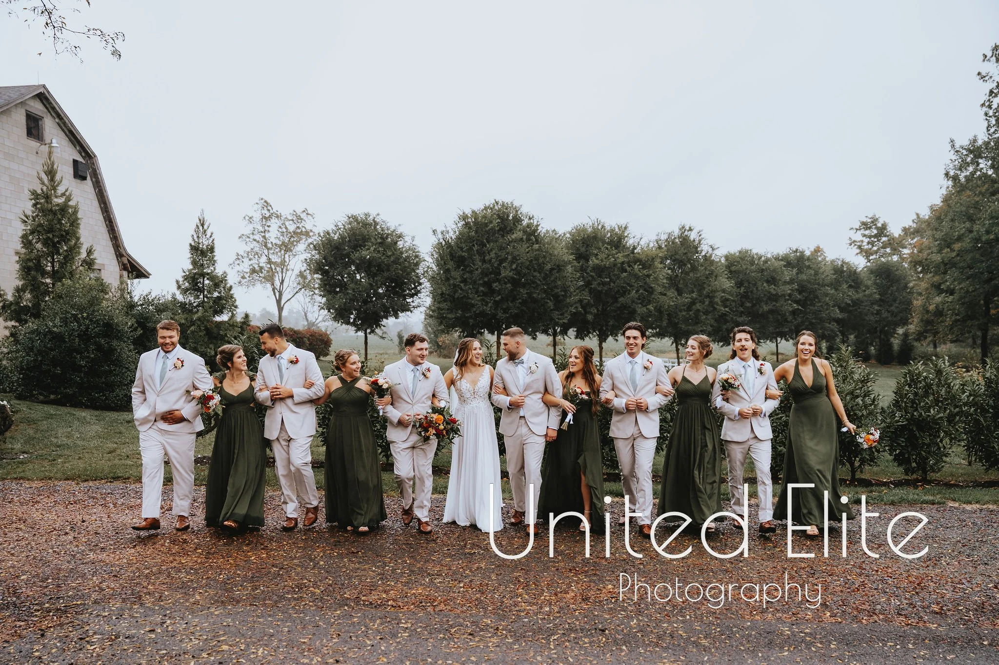 A group of wedding party members walking outdoors on a cloudy day, with trees and a building in the background.