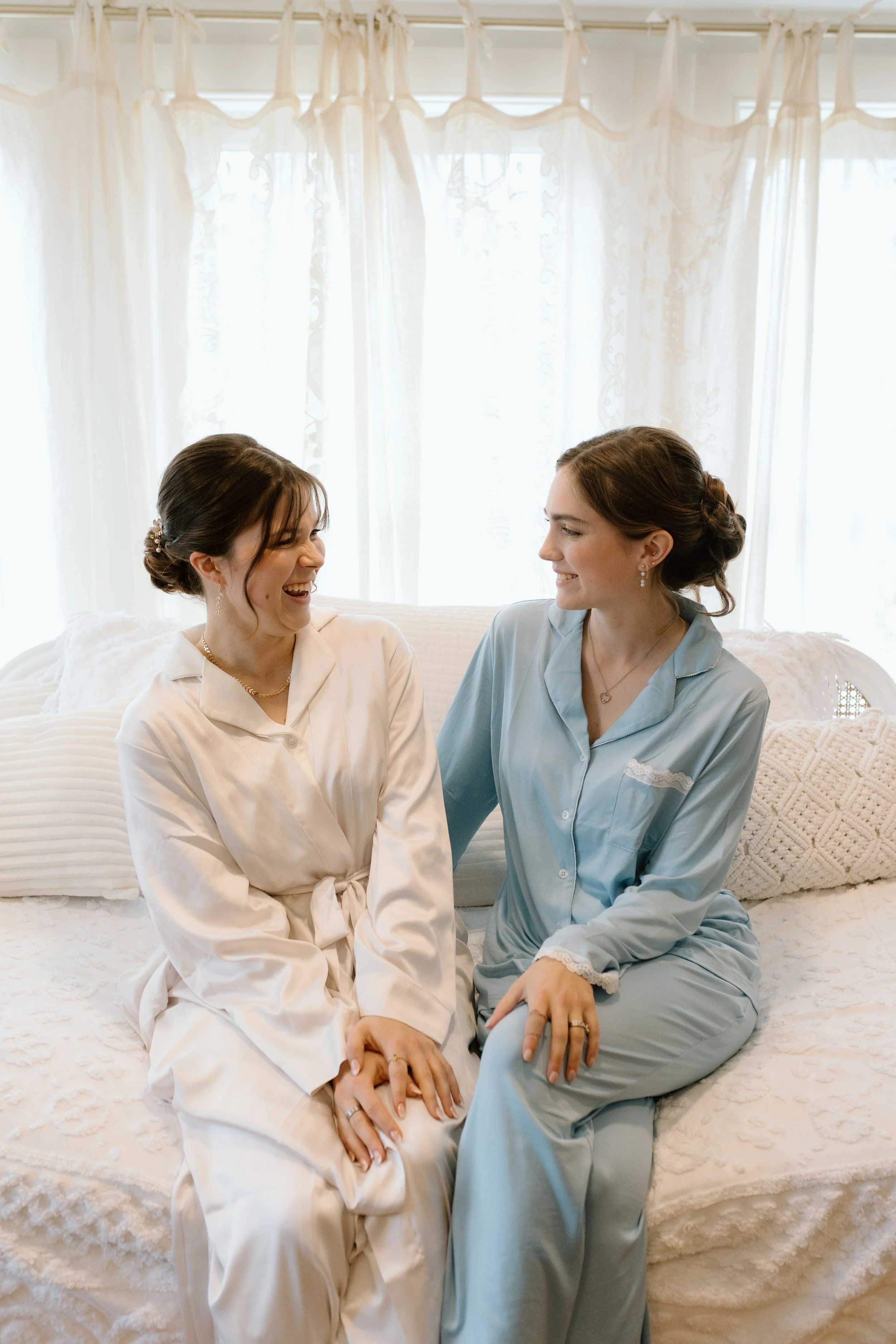 Two women sitting on a bed, facing each other and smiling. One woman is wearing white pajamas, and the other is wearing light blue pajamas. They are holding hands and seem to be sharing a happy moment in a bright room with sheer white curtains.