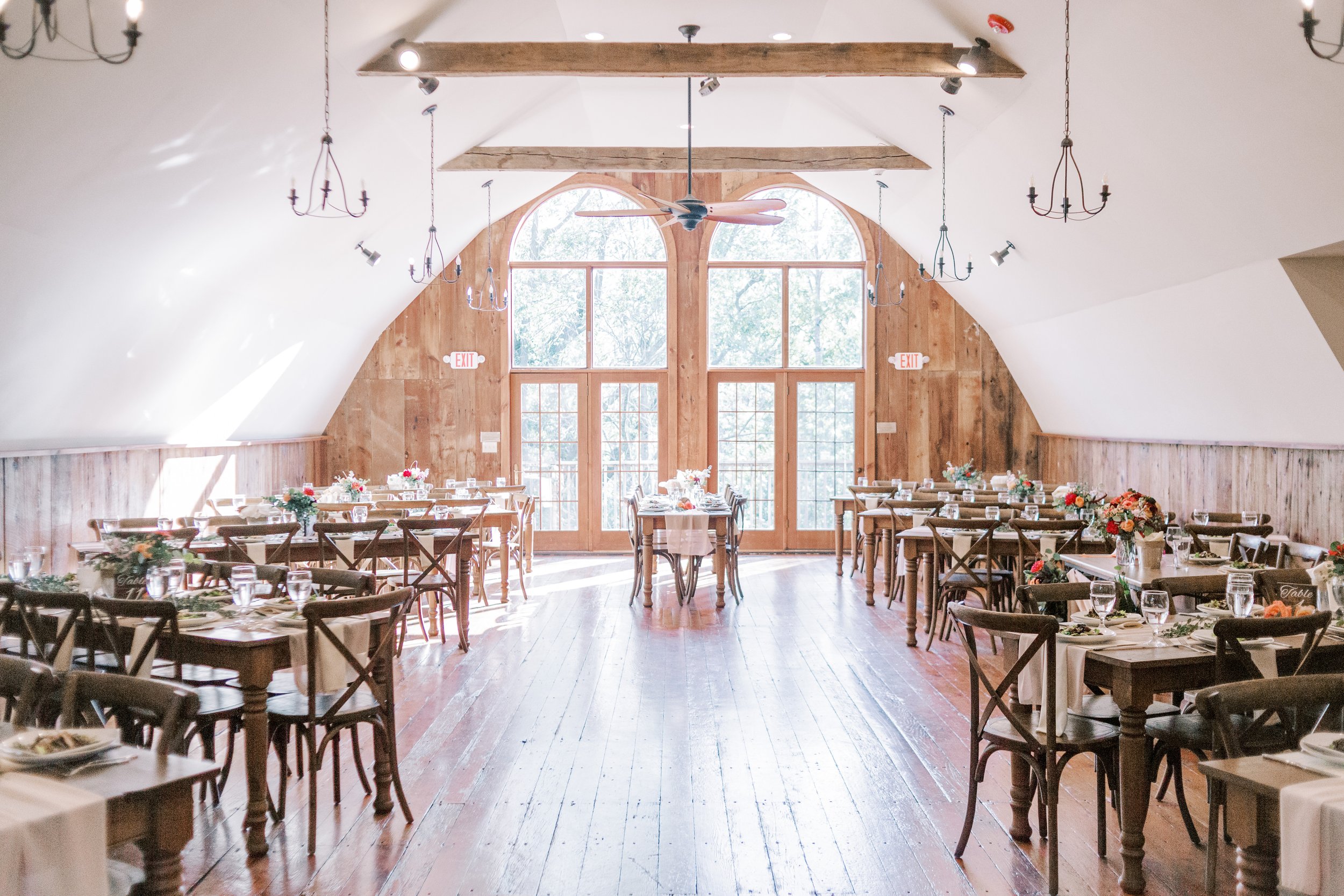 A spacious banquet hall with wooden flooring, high vaulted white ceiling with wooden beams, large glass windows with arched tops, and wooden walls. Multiple tables are set with white tablecloths, floral centerpieces, and glassware, arranged for a for