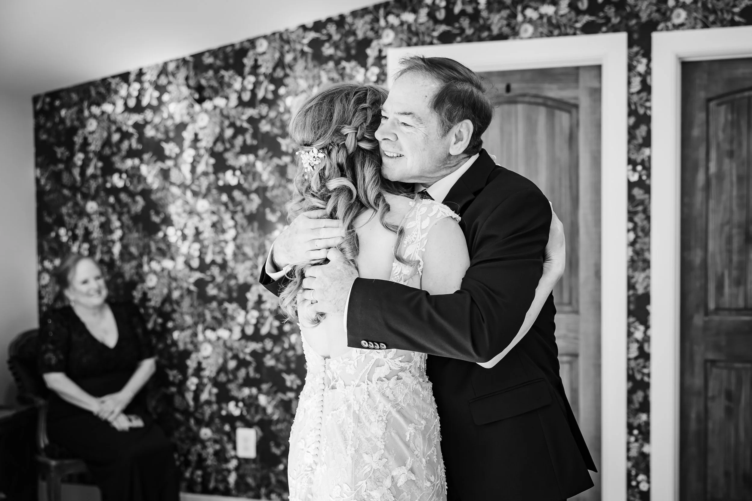 A bride and groom embrace during their wedding ceremony, smiling at each other. The bride wears a lace wedding dress with floral details in her hair, while the groom is in a dark suit. An attentive woman, possibly a guest or officiant, sits in the ba