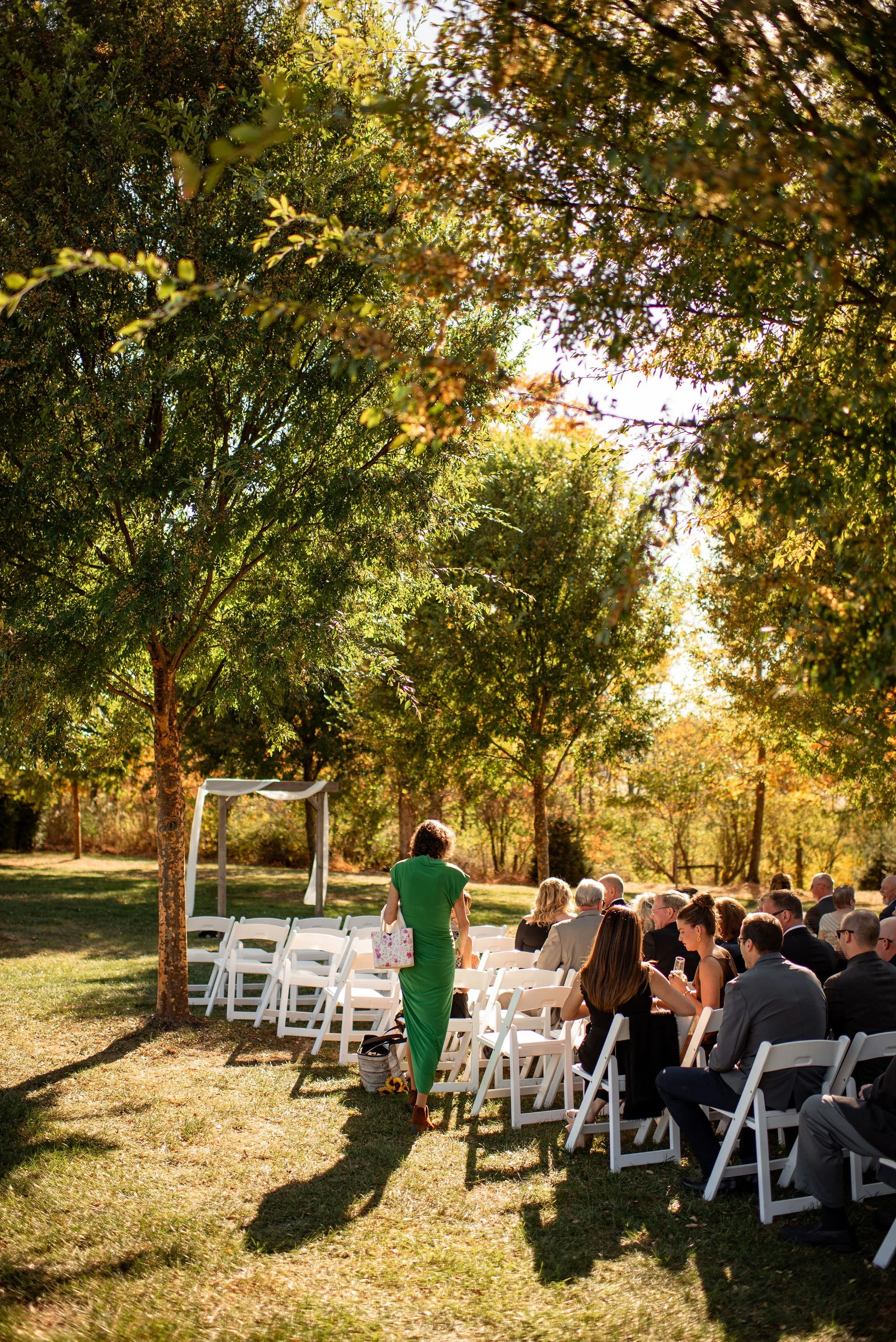 An outdoor wedding ceremony set up in a park with white chairs arranged in rows and a white arch decorated with fabric at the front. A woman in a green dress is walking among the seated guests, who are dressed formally. There are tall, leafy trees pr