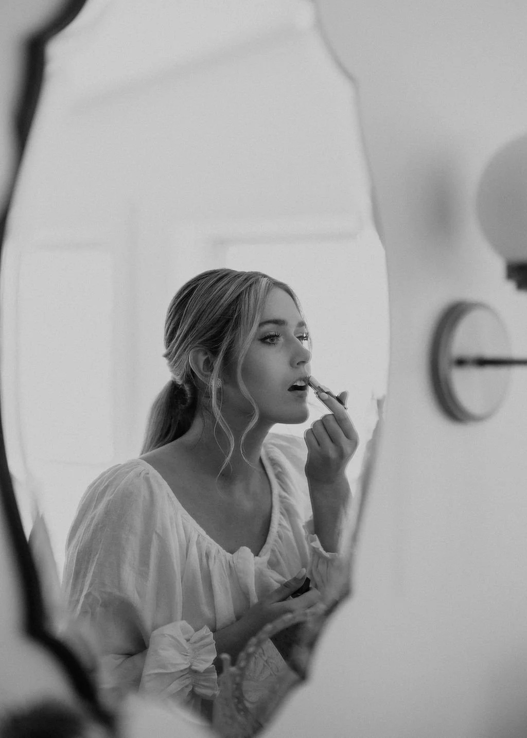 A woman with blonde hair styled in loose waves is seen applying lipstick, reflected in an irregularly shaped mirror. She is wearing a light-colored, puffed-sleeve top and has a calm, focused expression.