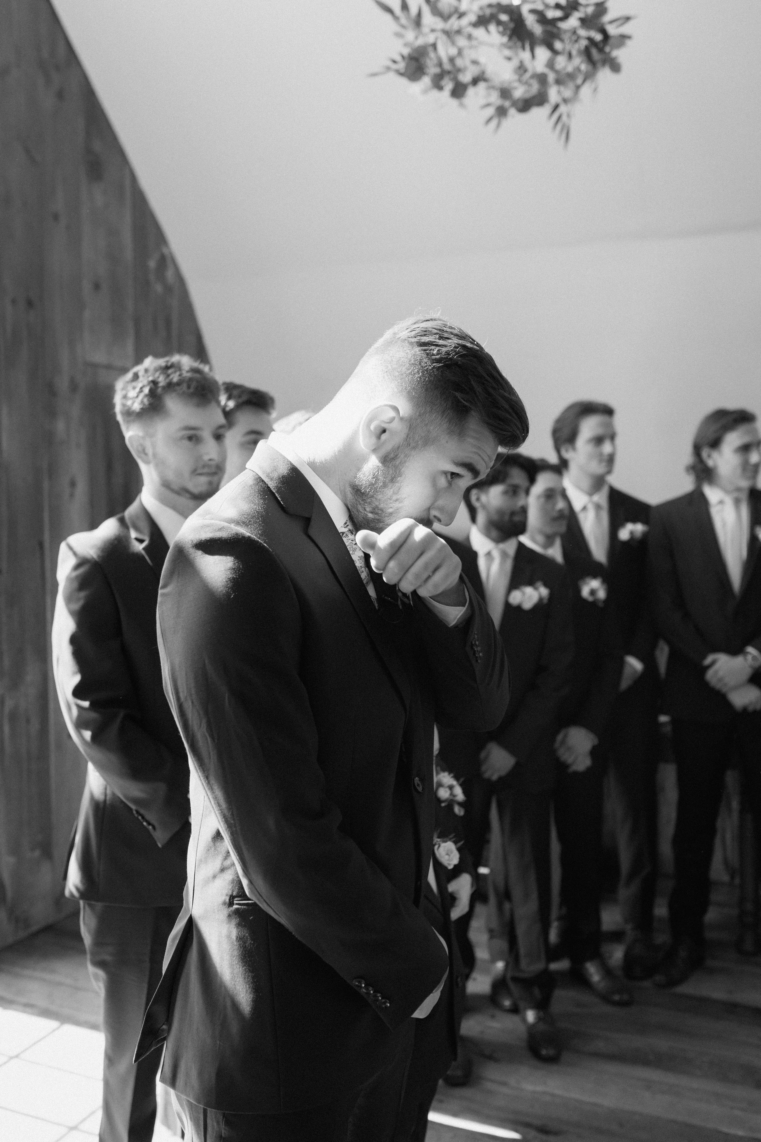 Black and white photo of a groom surrounded by groomsmen, all dressed in suits, at a wedding ceremony. The groom is in the foreground, adjusting his cufflink or sleeve, with a serious or contemplative expression. The groomsmen stand in the background