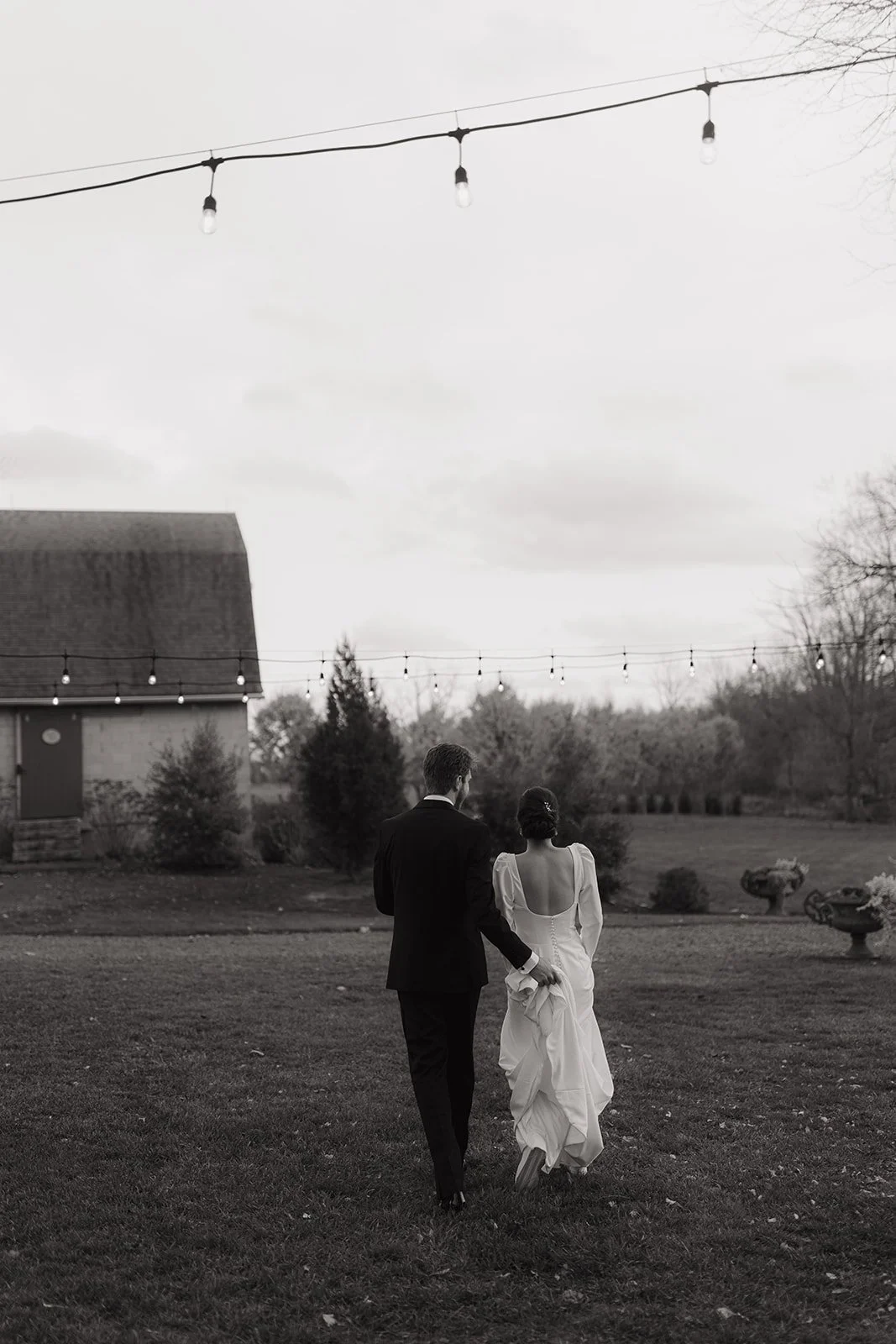 A black-and-white photo of a bride and groom walking hand in hand outdoors, with string lights hanging above them and a building with a roof and trees in the background.