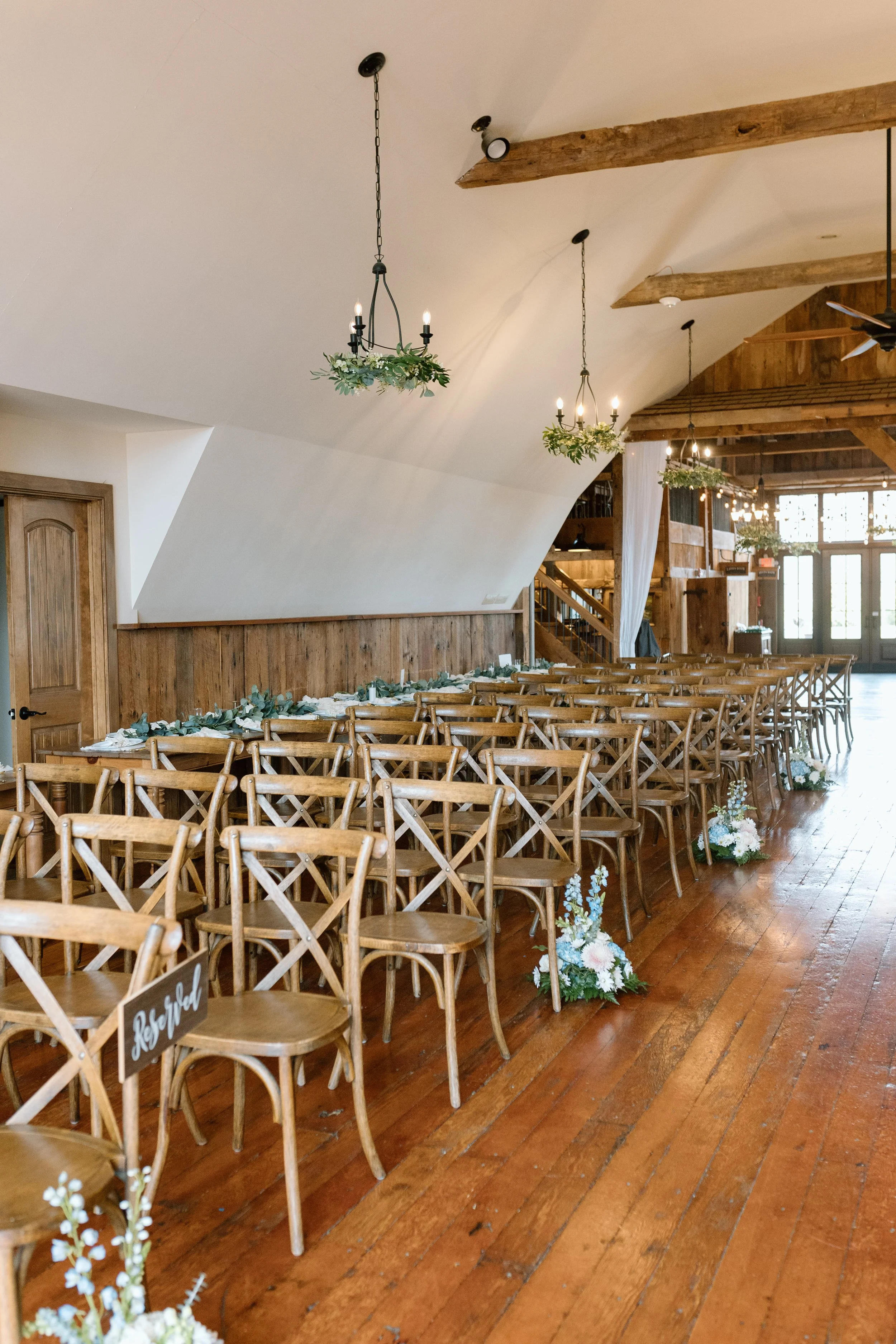 Indoor wedding ceremony setup with rows of wooden chairs decorated with white flowers and greenery, large chandeliers with greenery hanging from the ceiling, and rustic wooden beams in a warmly lit space.