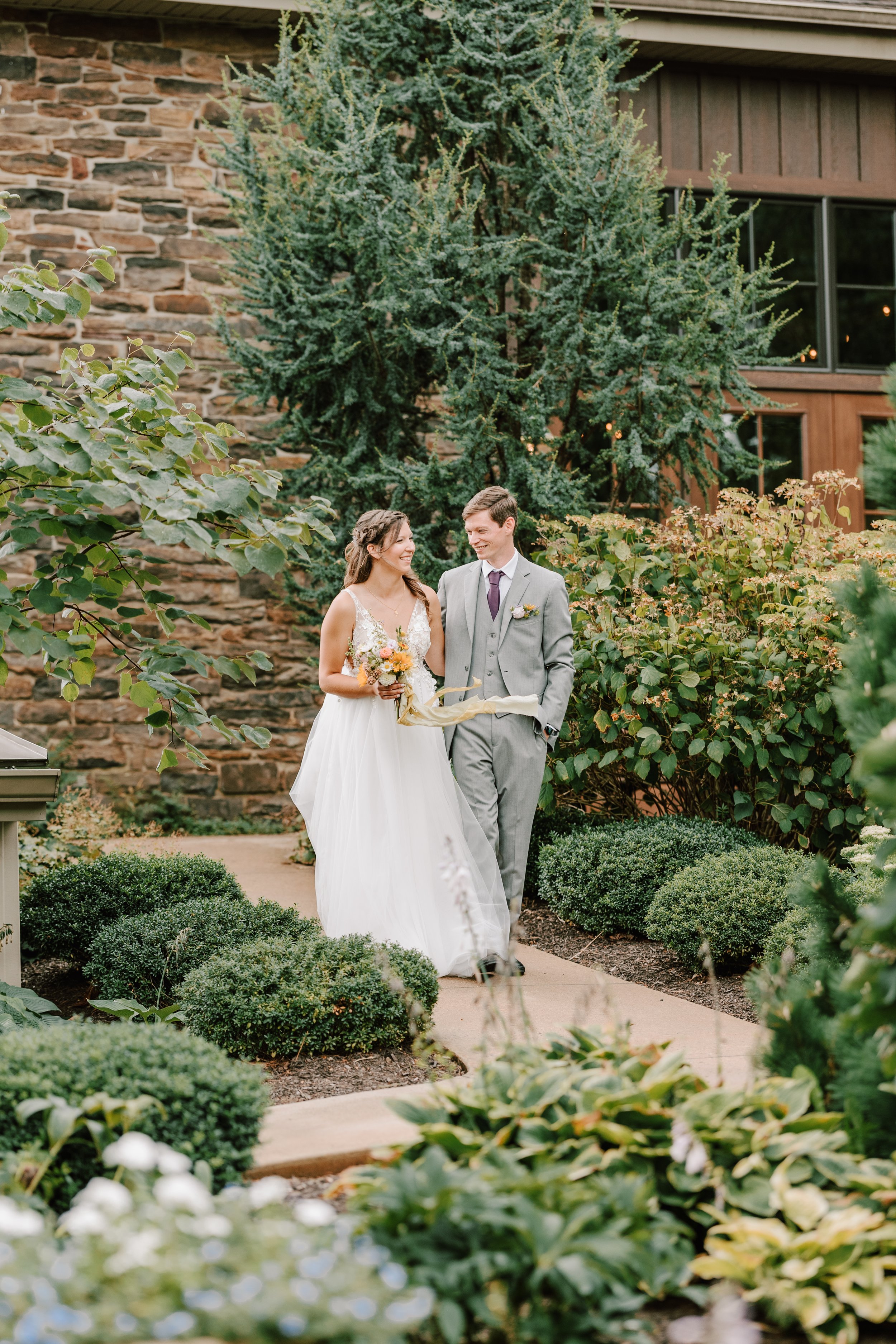 A bride and groom walking hand in hand on a garden pathway during a wedding ceremony. The bride is wearing a white dress and holding a bouquet of flowers, while the groom is dressed in a gray suit with a boutonniere.
