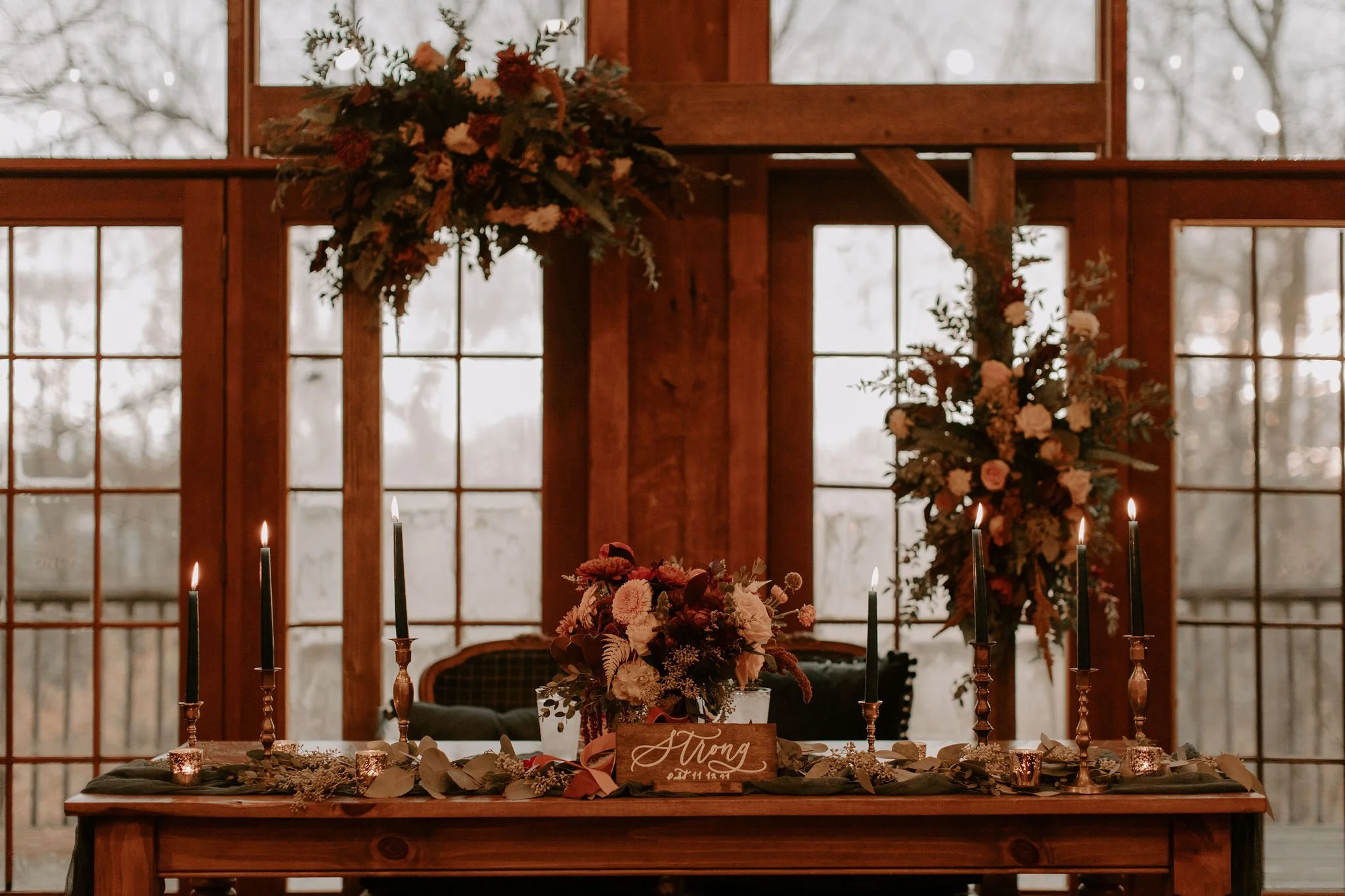 Decorated table with flowers, candles, and candlescapes, set in front of large window with wooden framing.