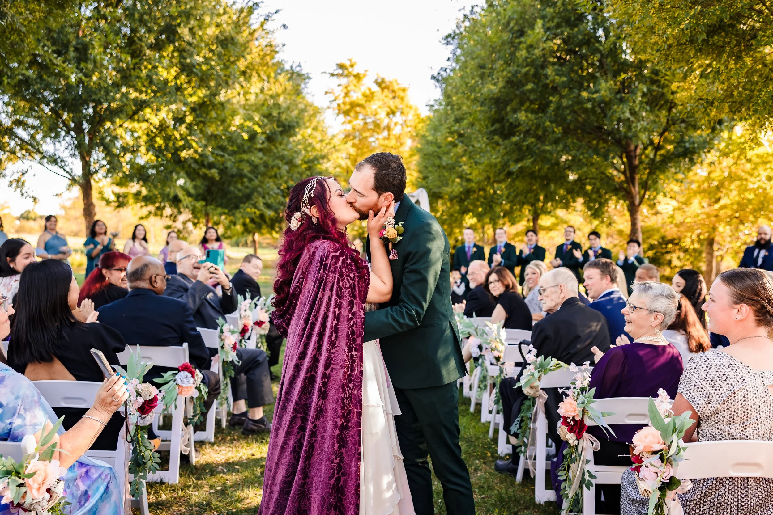 A couple kisses during an outdoor wedding ceremony surrounded by seated guests and lush green trees, with sunlight filtering through the leaves.