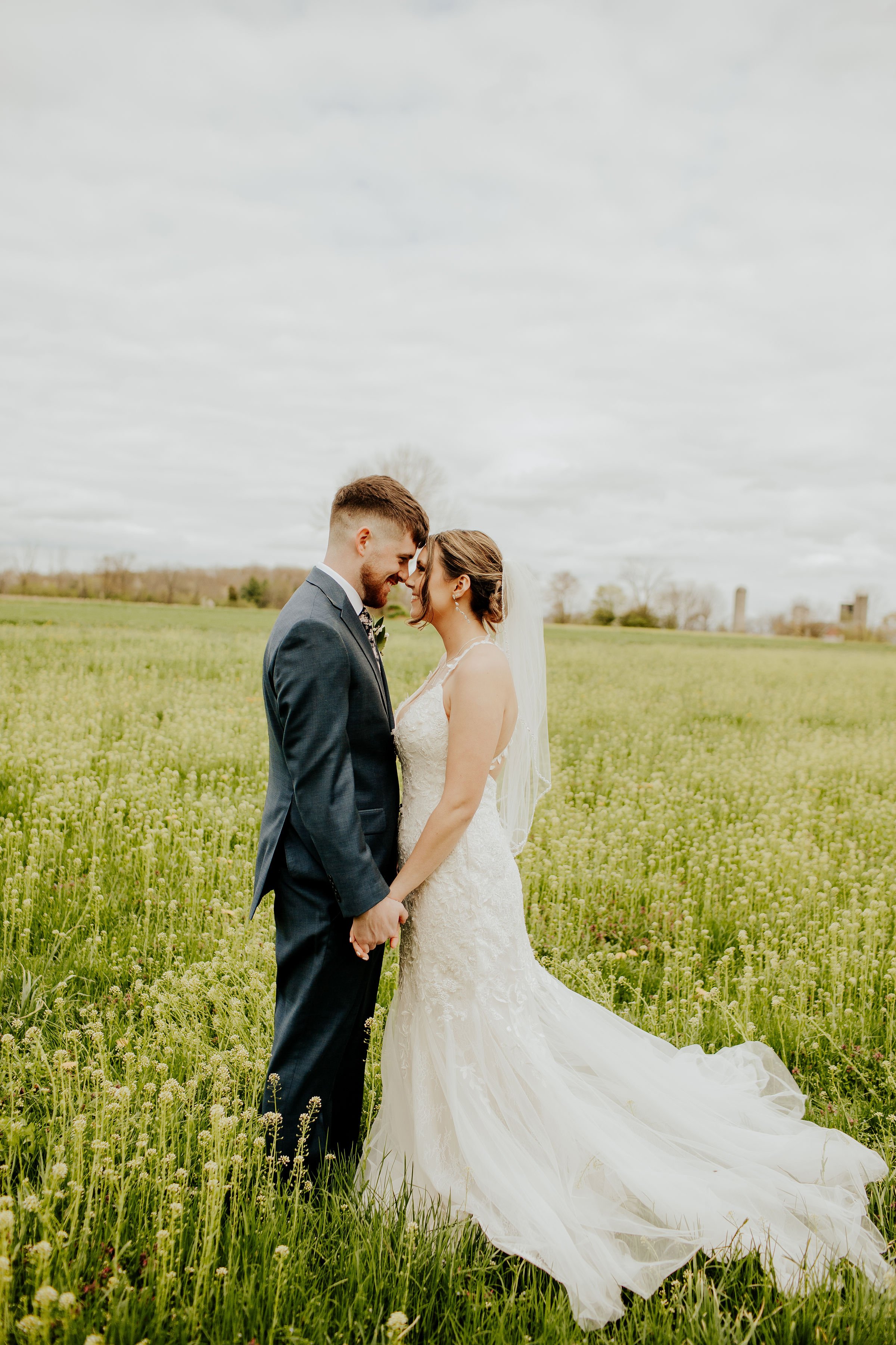 A bride and groom in wedding attire holding hands in a grassy field, close to each other with their foreheads touching, during daytime with cloudy sky.