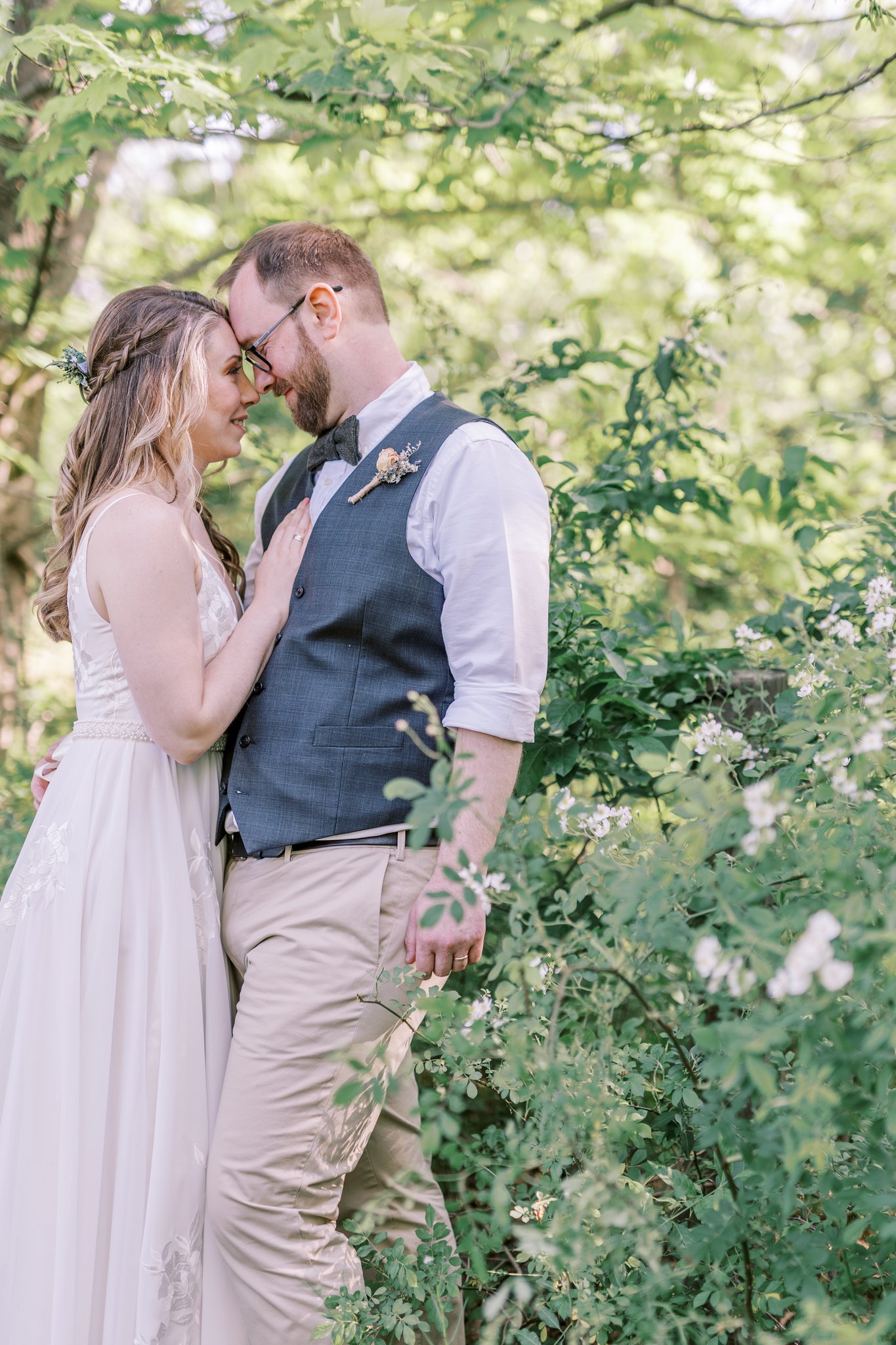 A couple dressed in wedding attire standing close with foreheads touching in a lush green outdoor setting surrounded by trees and flowering bushes.