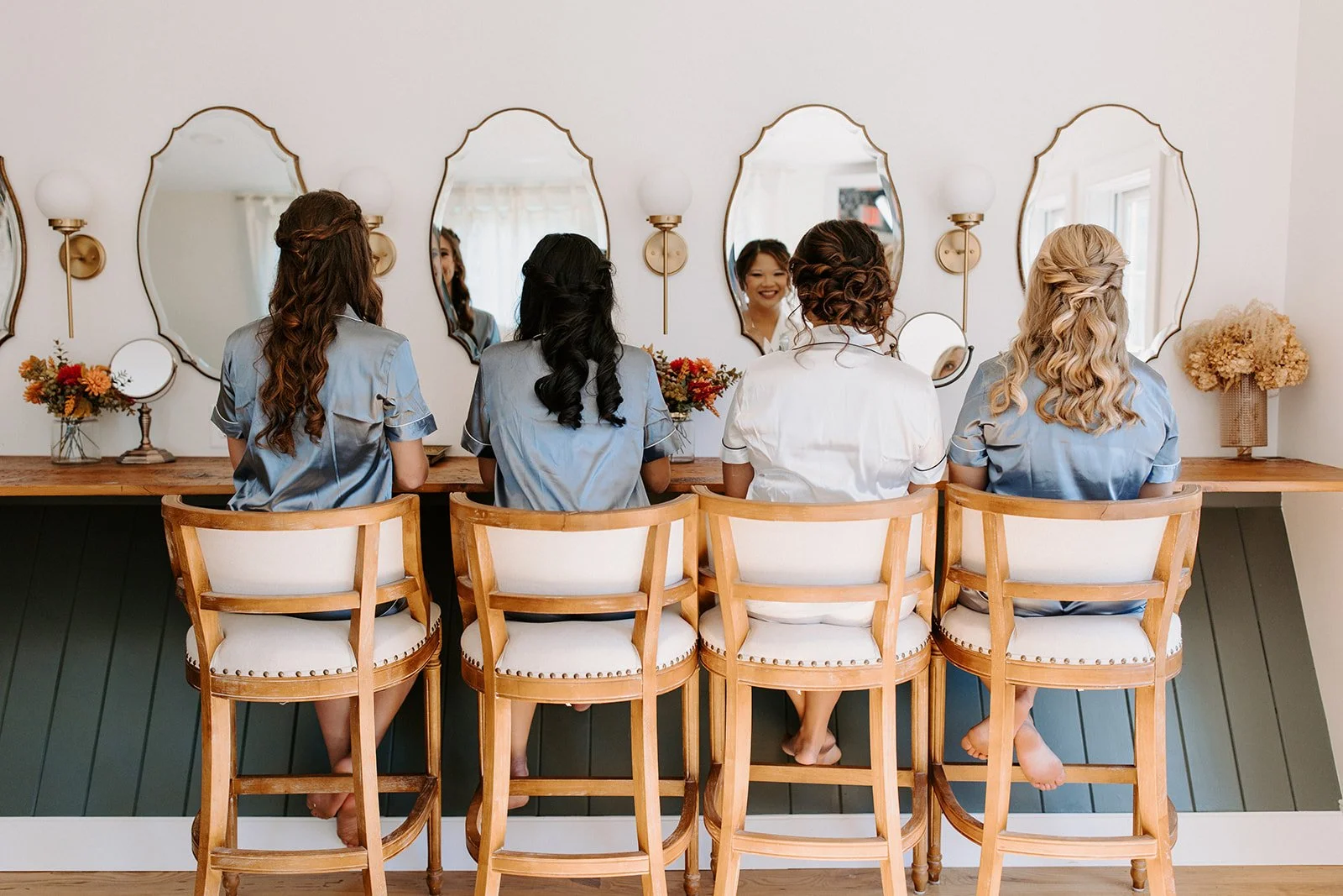 Four women sitting at a vanity table with mirrors, getting ready, wearing matching blue satin pajamas, with one woman smiling at her reflection.