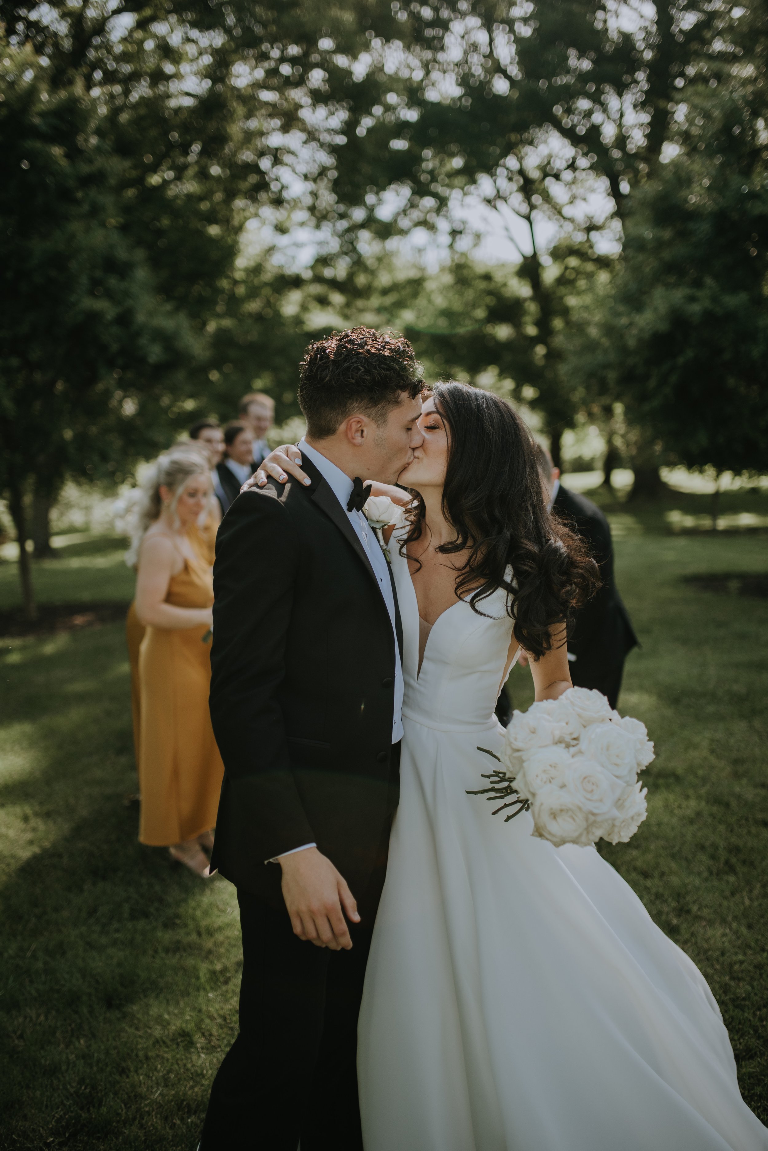 A bride and groom kiss outdoors at their wedding, with bridesmaids and groomsmen in the background under green trees.