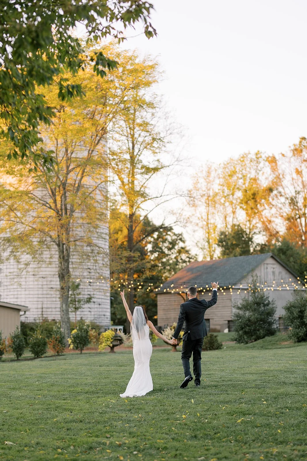 A bride and groom holding hands and walking across a grassy area during sunset, with trees and a rustic barn in the background.