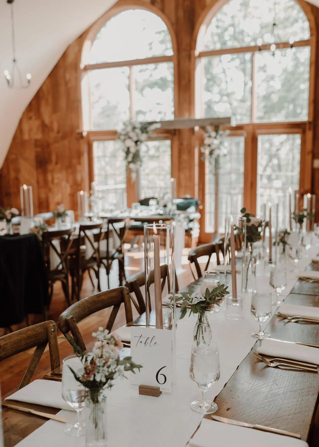 Indoor wedding reception setup with wooden tables, chairs, floral centerpieces, and tall candleholders in a rustic room with large arched windows and wood paneling.