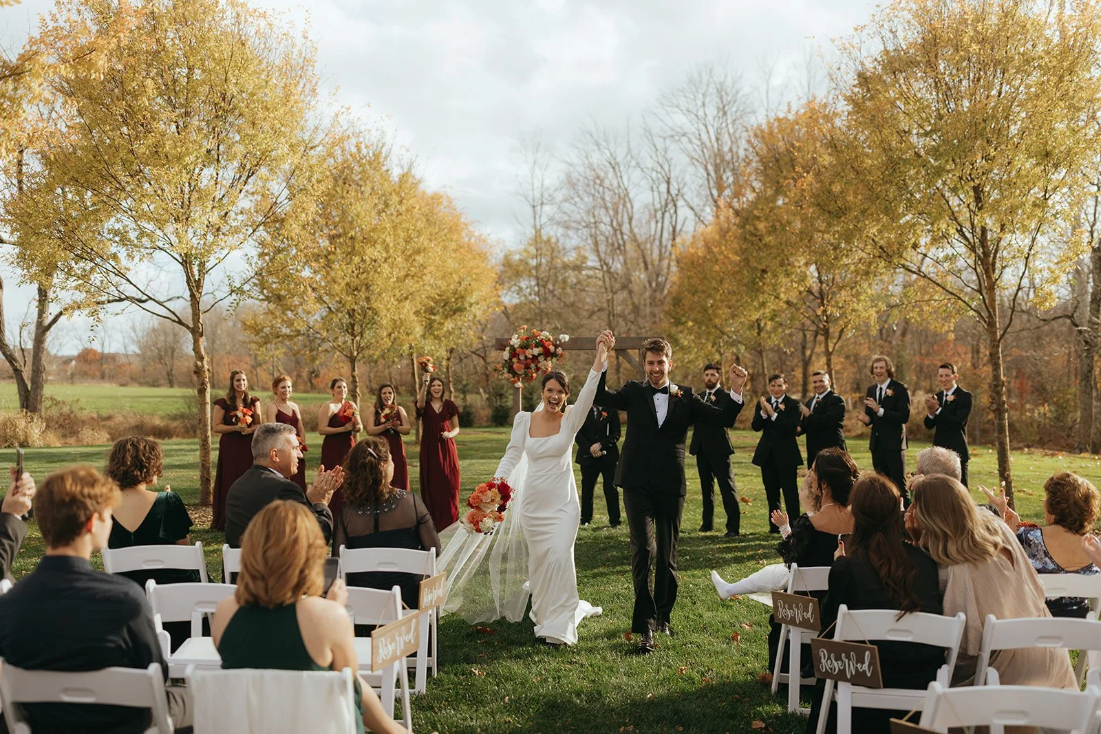 A newlywed couple celebrates their wedding outdoors in a park during autumn, walking hand in hand amidst  guests seated on white chairs and standing, with trees showing yellow and orange leaves in the background.