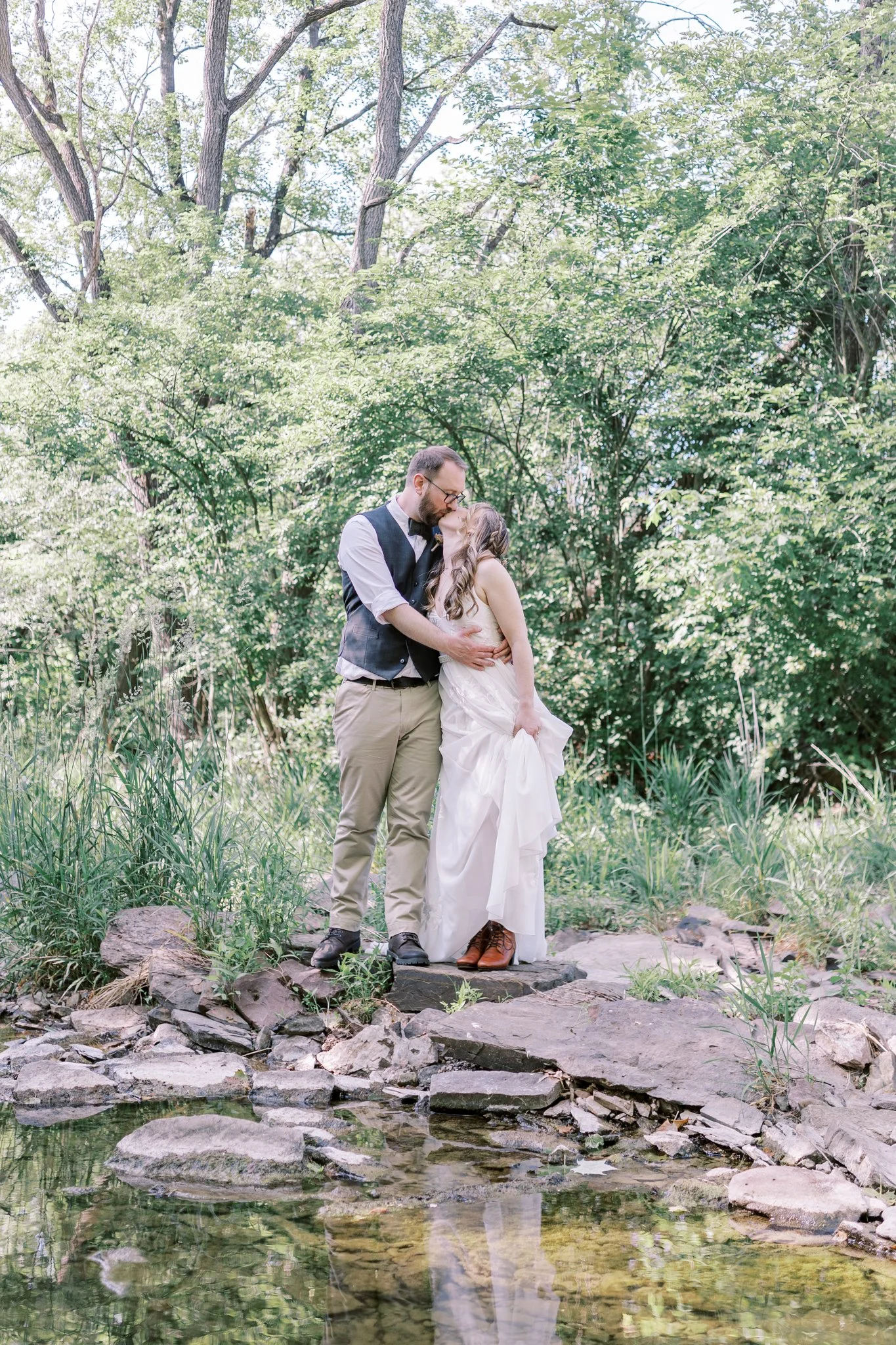 A couple kissing outdoors on a rocky path beside a small stream, surrounded by lush green trees and foliage.