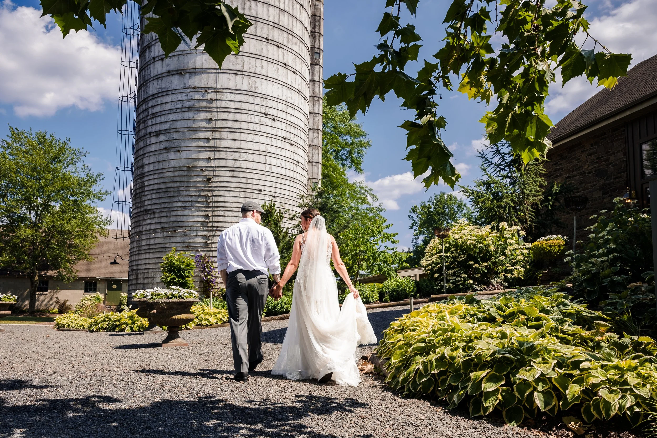 A bride and groom holding hands during their outdoor wedding ceremony in a lush garden, with a large silo and a building in the background, under a blue sky with scattered clouds.