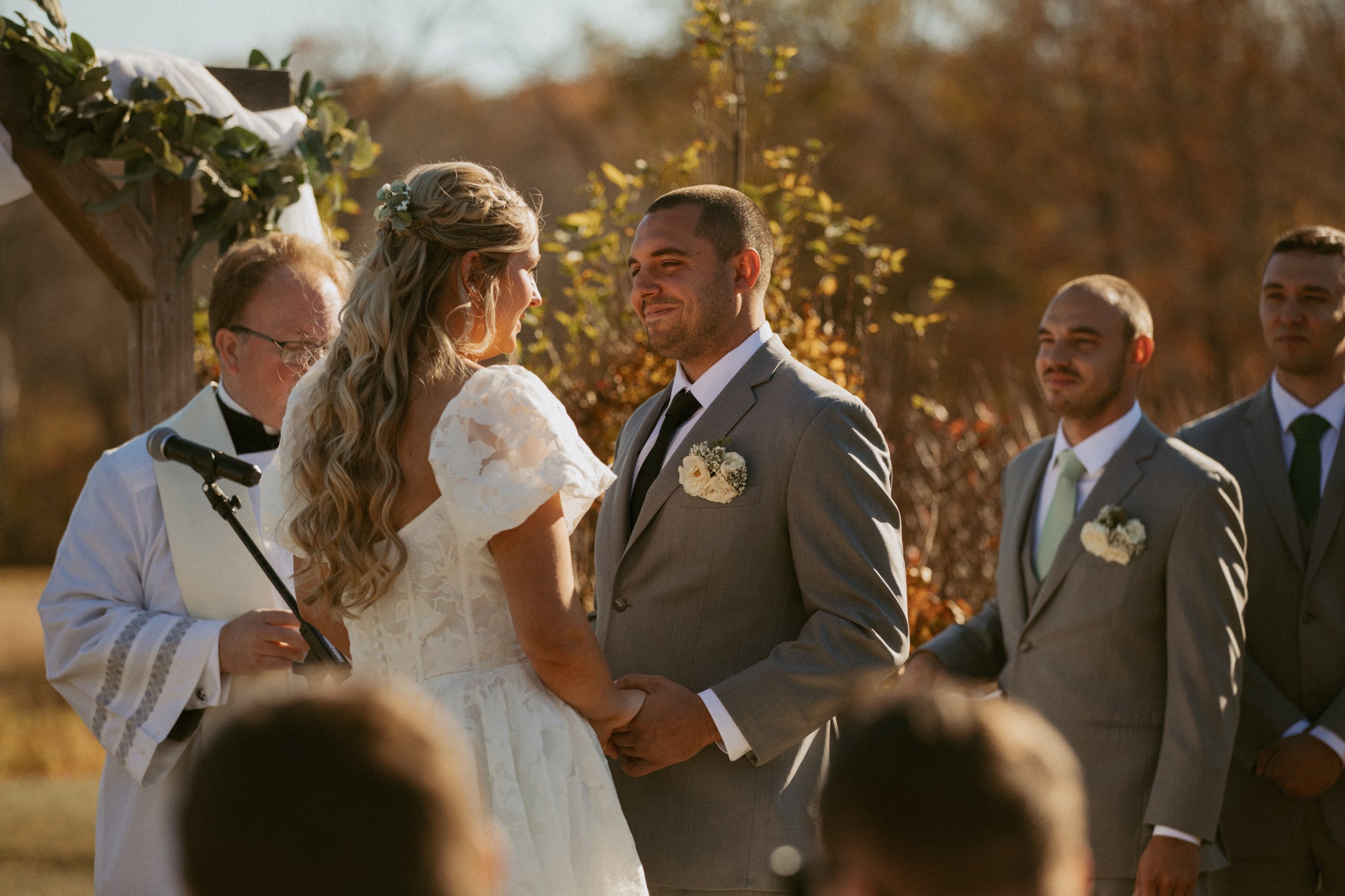 A couple exchanges vows outdoors at a wedding, surrounded by their wedding party dressed in gray suits, with autumn trees in the background.