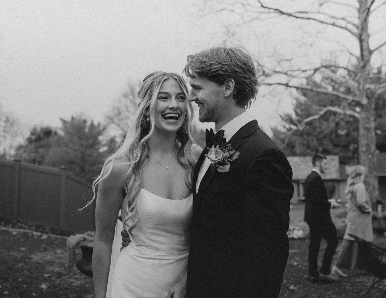 A black and white photo of a bride and groom smiling at each other at their wedding, outdoors with bare trees and other wedding guests in the background.