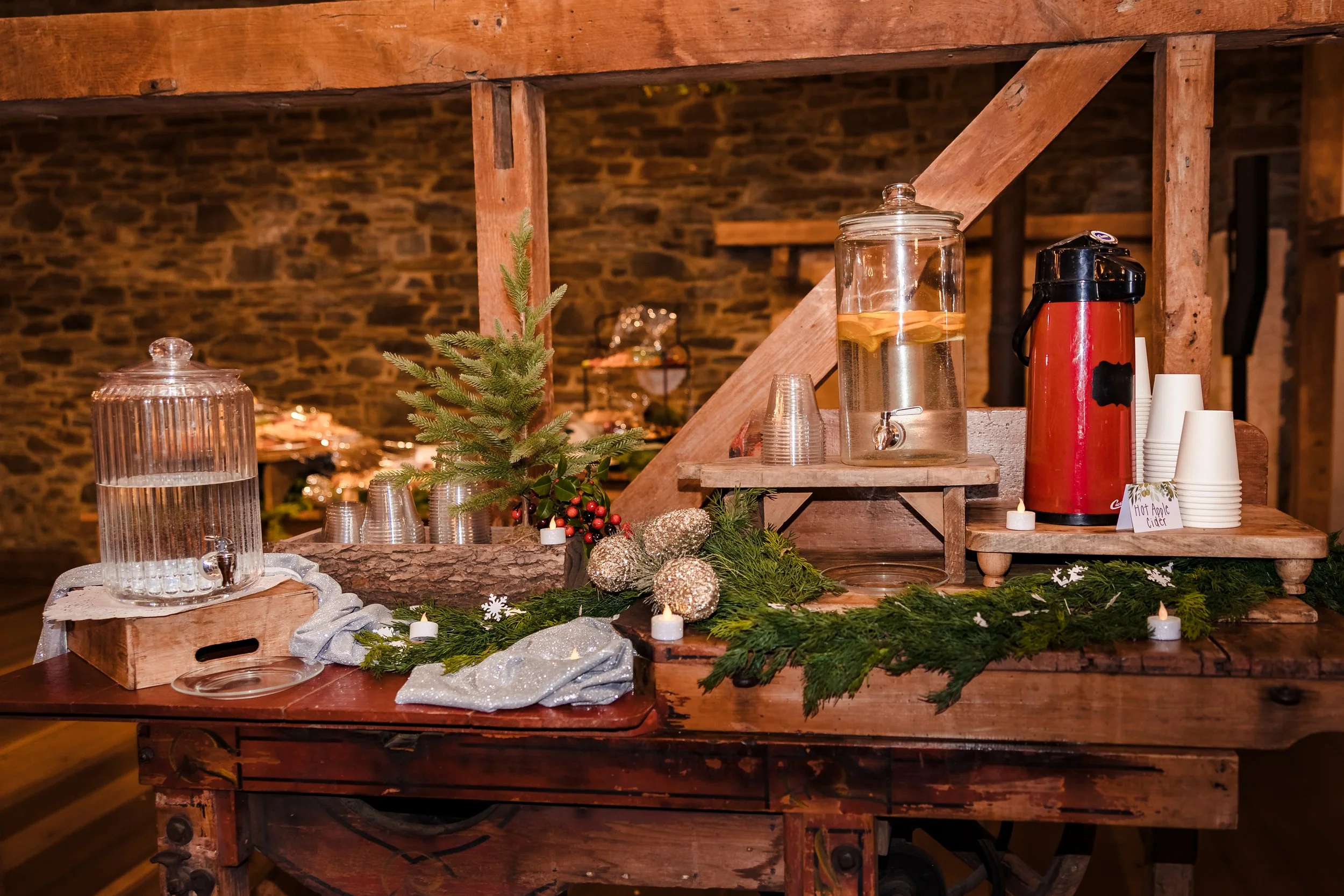 A rustic wooden table decorated for the holidays with a small pine tree, candles, silver ornaments, greenery, and a beverage station featuring a glass jar of water with lemon slices, a red drink dispenser, cups, and a sign reading 'Hot Apple Cider'.