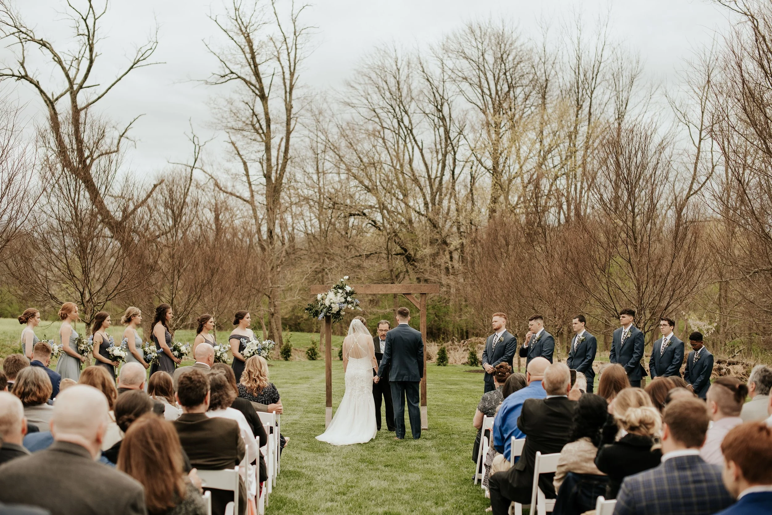 Outdoor wedding ceremony with the bride and groom standing before an officiant, surrounded by bridesmaids and groomsmen, with an audience seated on white chairs on a grassy field and leafless trees in the background.