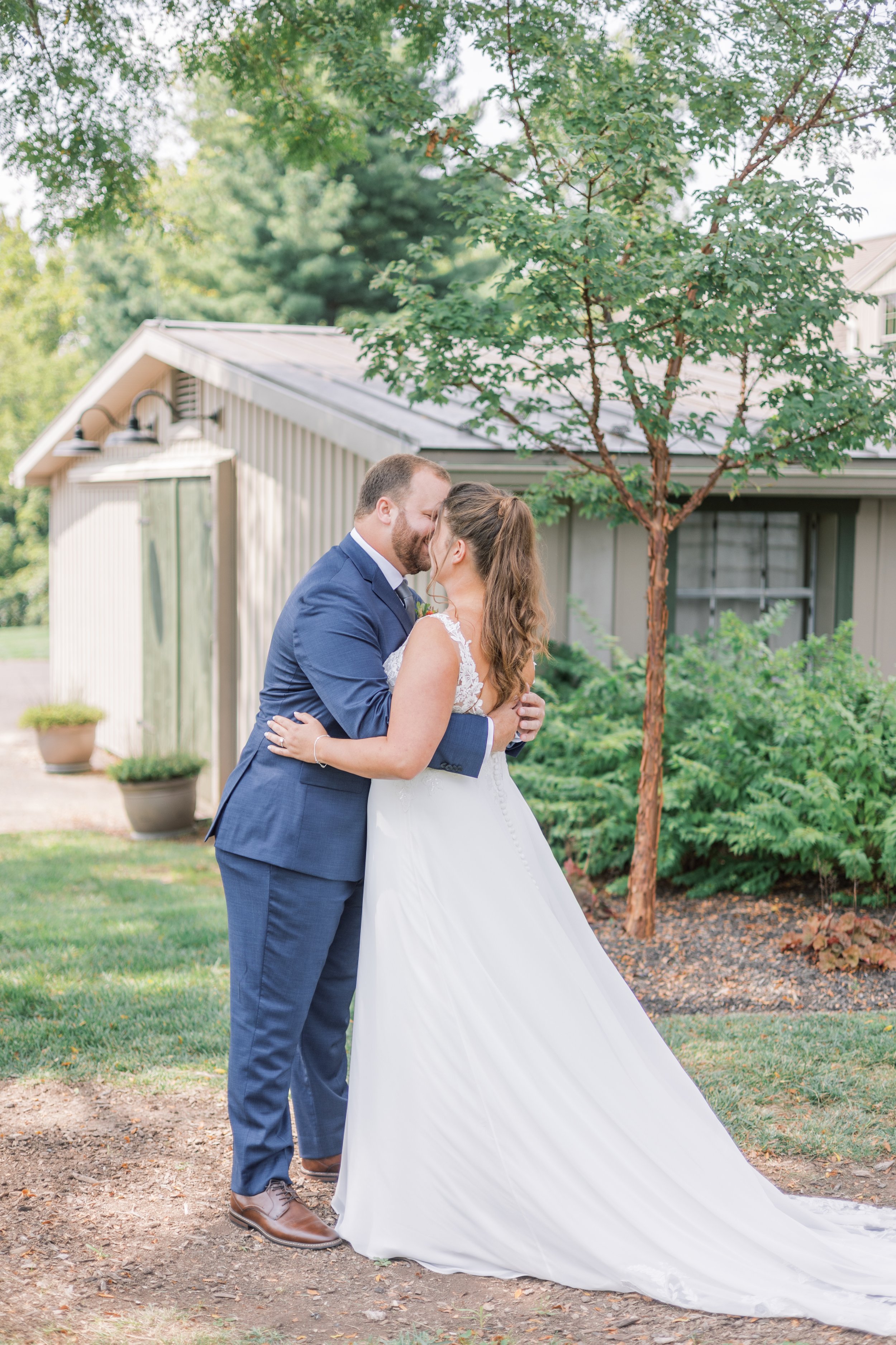 A bride and groom kissing outdoors near a tree and house, wedding dress and suit.