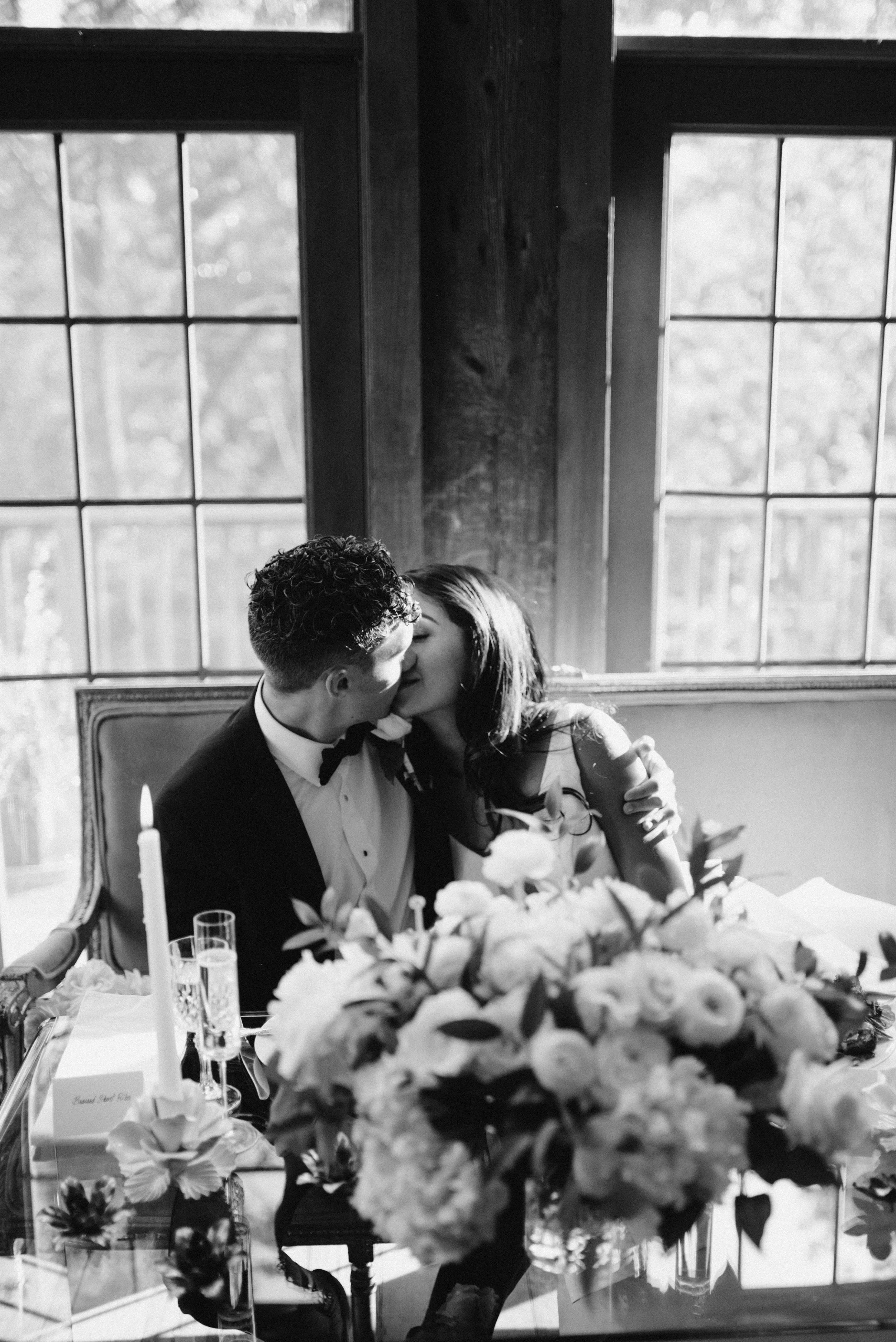 A couple sharing a kiss at a wedding reception with floral arrangements and candles on the table.