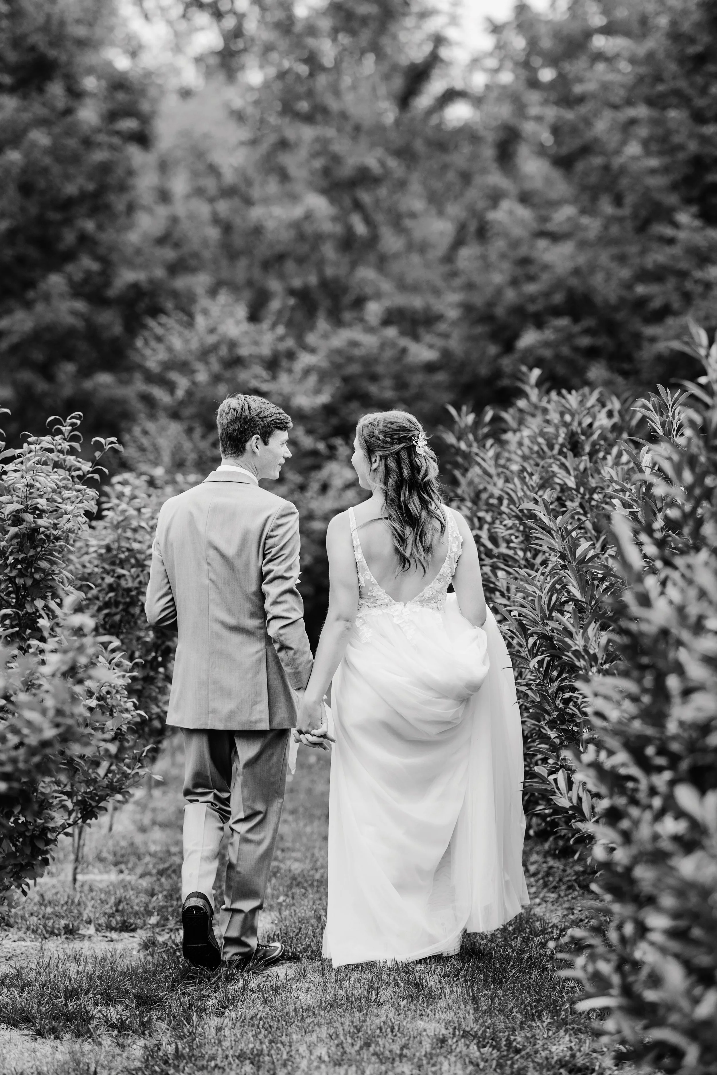 A black and white photograph of a bride and groom holding hands and walking in a garden, surrounded by bushes and trees.