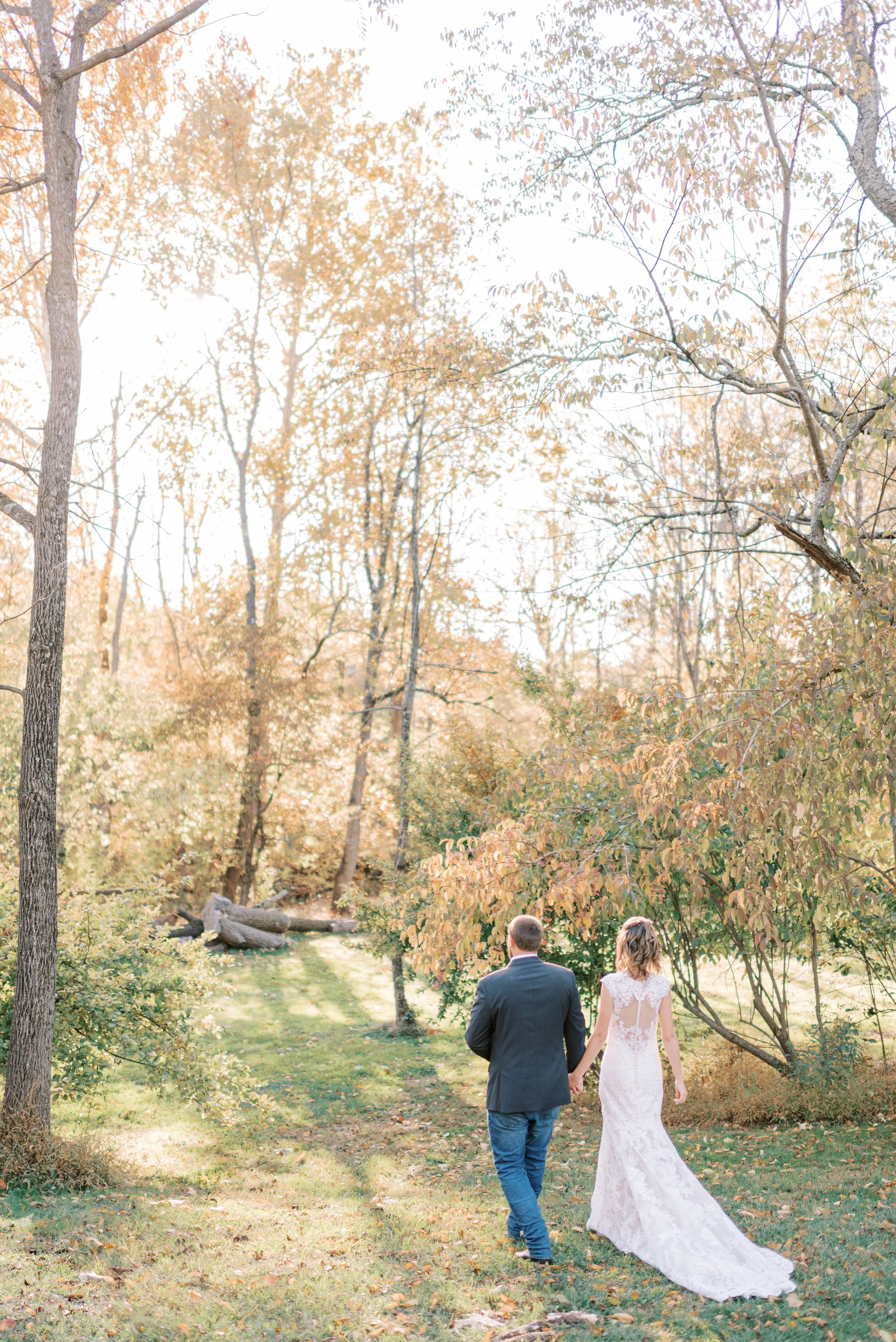A newlywed couple, a man in a suit and a woman in a white lace wedding dress, walk hand in hand through a wooded park during autumn.