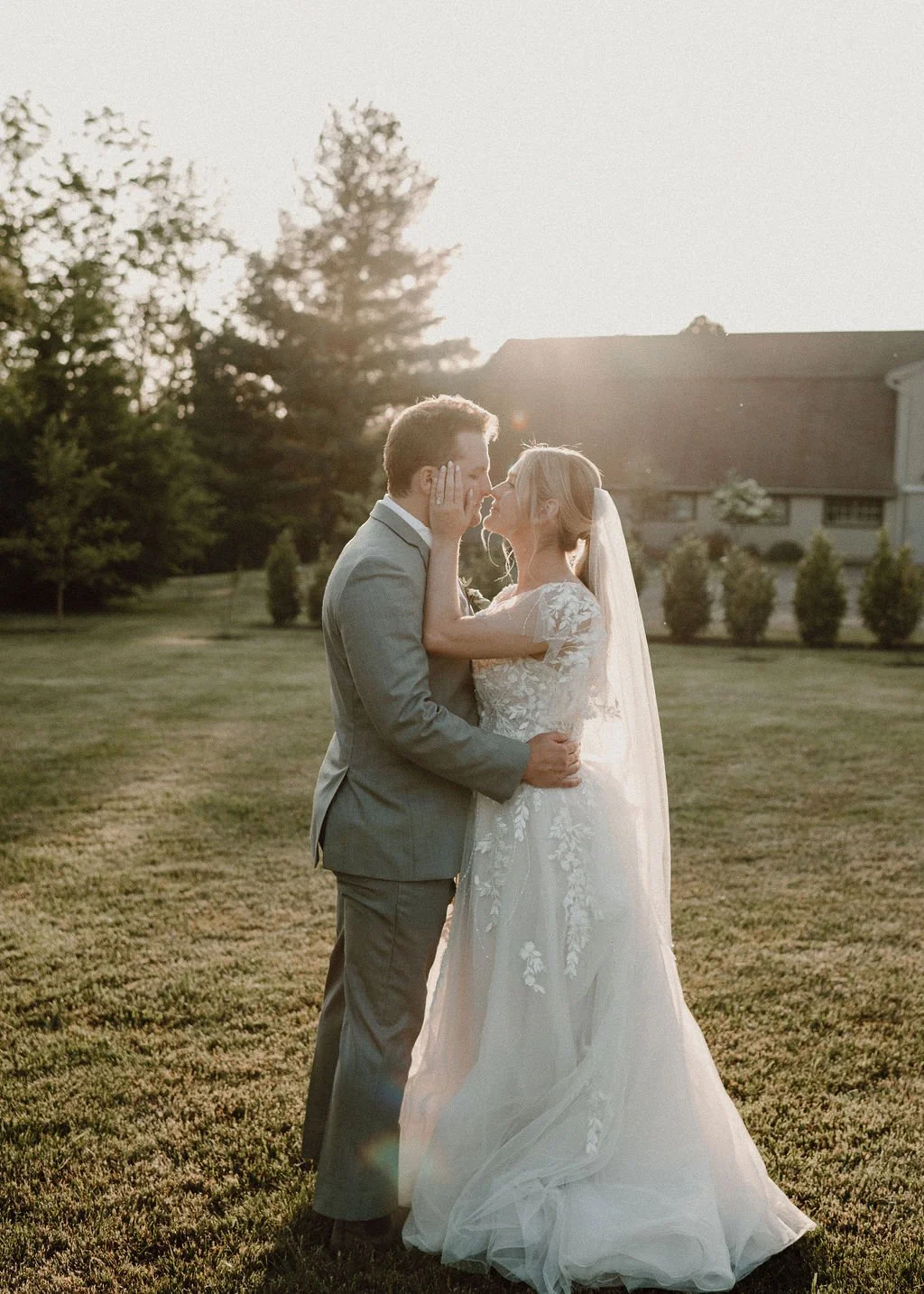 A bride and groom share a romantic moment outdoors at sunset, standing on a grassy lawn with trees and a house in the background.