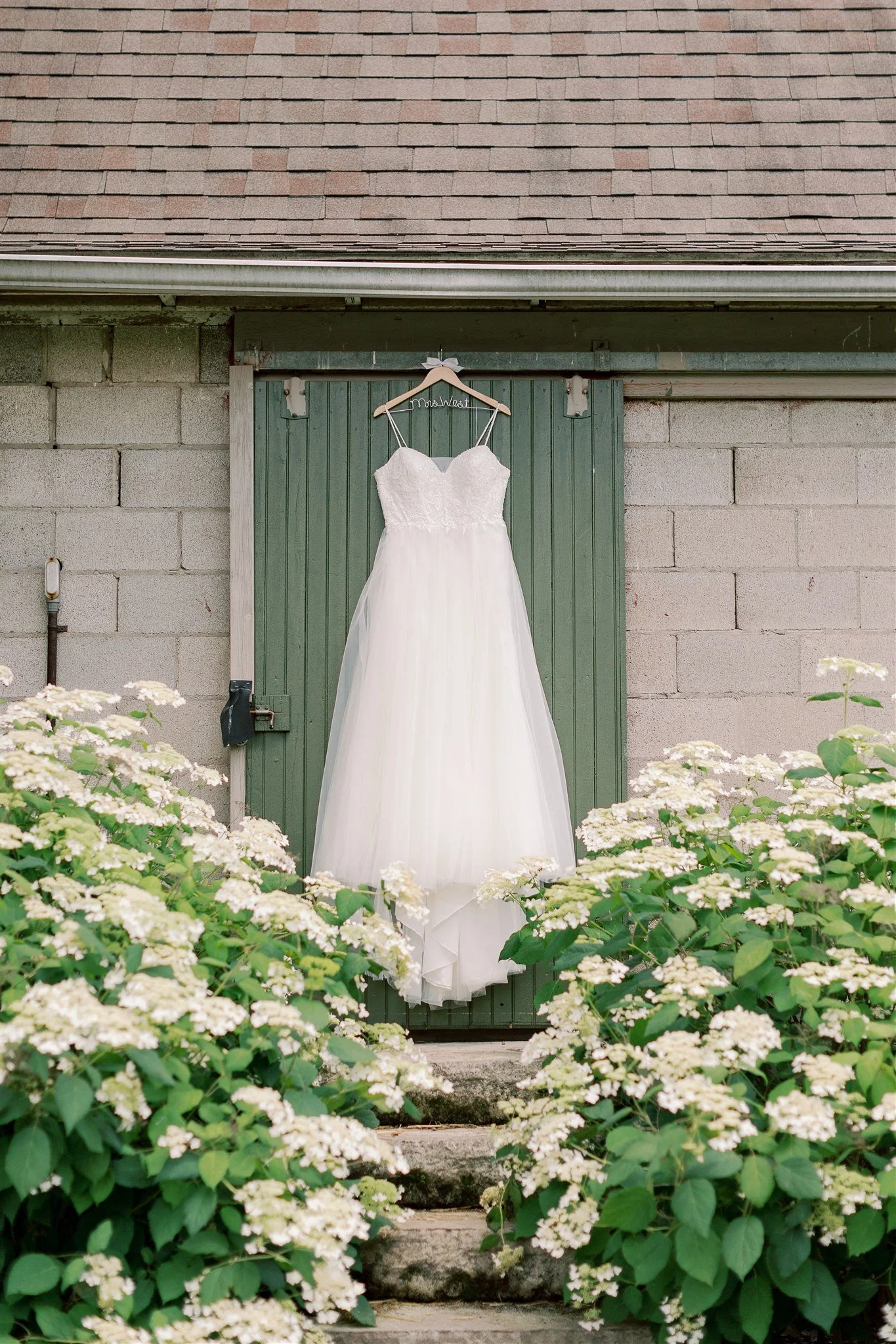 White wedding dress hanging on a tree branch in front of a green door, surrounded by white flowering plants.