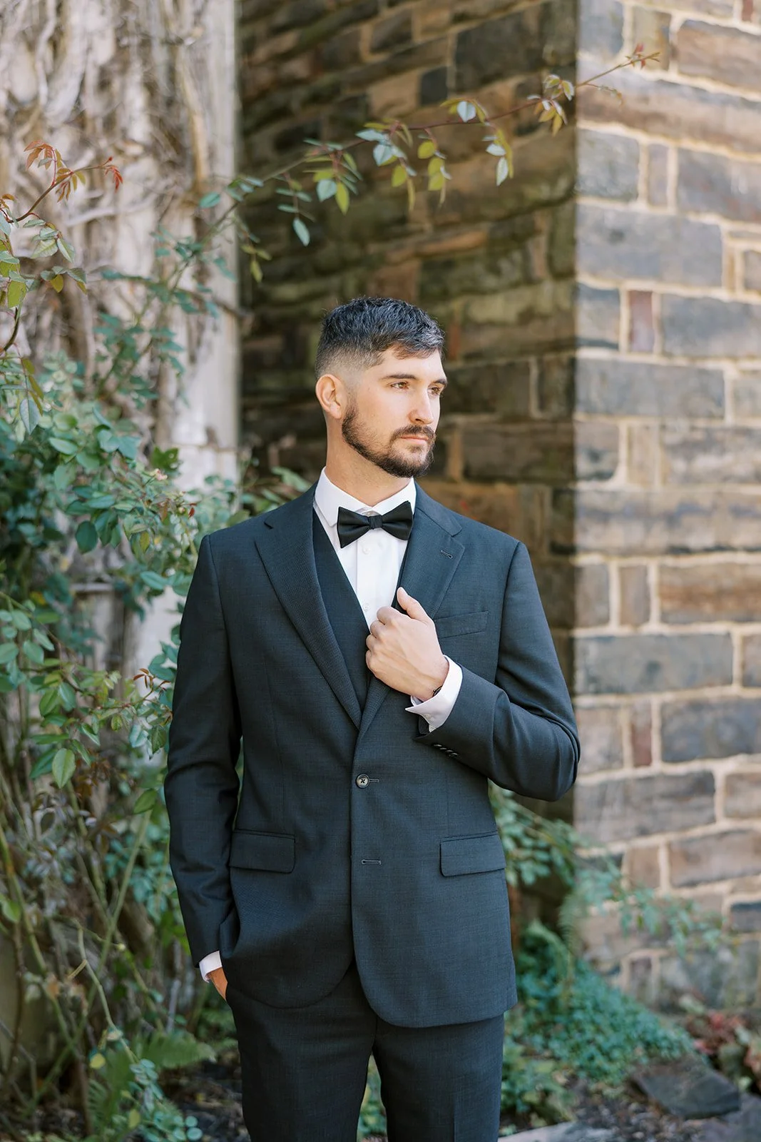 A man in a black tuxedo with a bow tie standing outdoors next to a brick wall and green foliage, looking thoughtfully to the side.