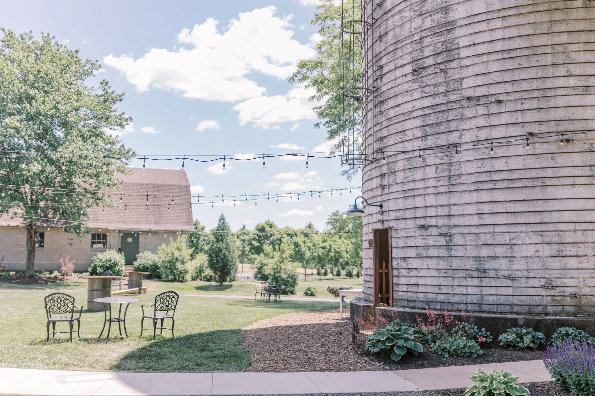 An outdoor seating area with a metal table and four chairs on a lawn. There are string lights hanging above, a large weathered silo on the right, trees, a building with a green door, and a bench in the background under a partly cloudy sky.