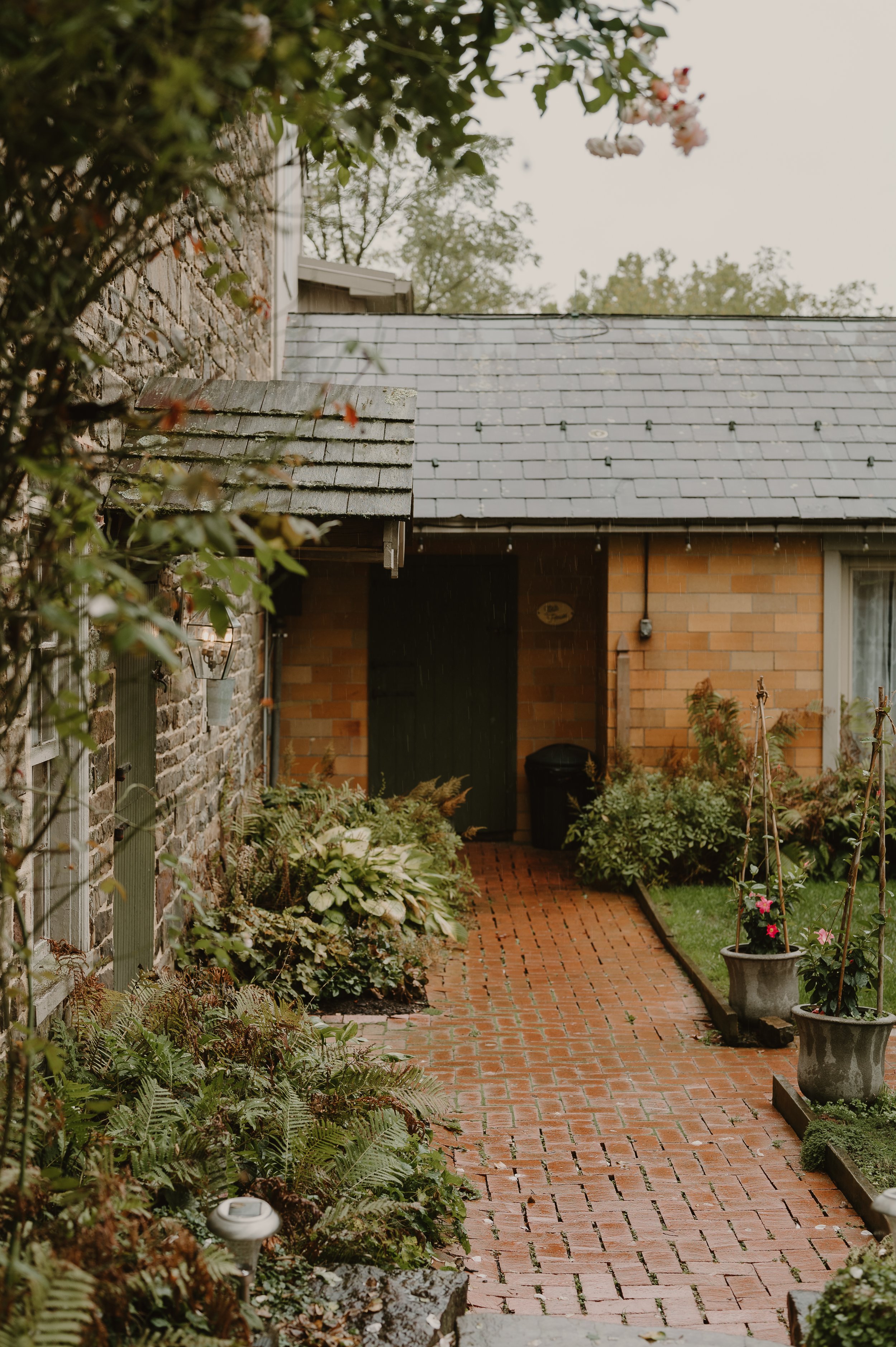 A brick pathway leading to the front door of a house, surrounded by lush plants and potted flowers, with a section of stone wall and the house's brick exterior visible.