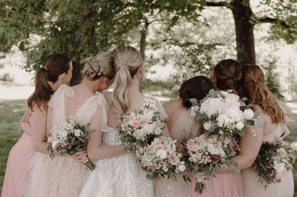 Group of women, likely bridesmaids, standing outdoors under trees, holding bouquets of flowers and wearing pastel-colored dresses.