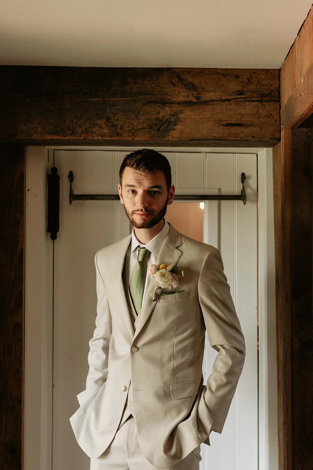 Young groom in a cream-colored suit with a green tie and boutonniere, standing in front of a mirror and posing for a photo.