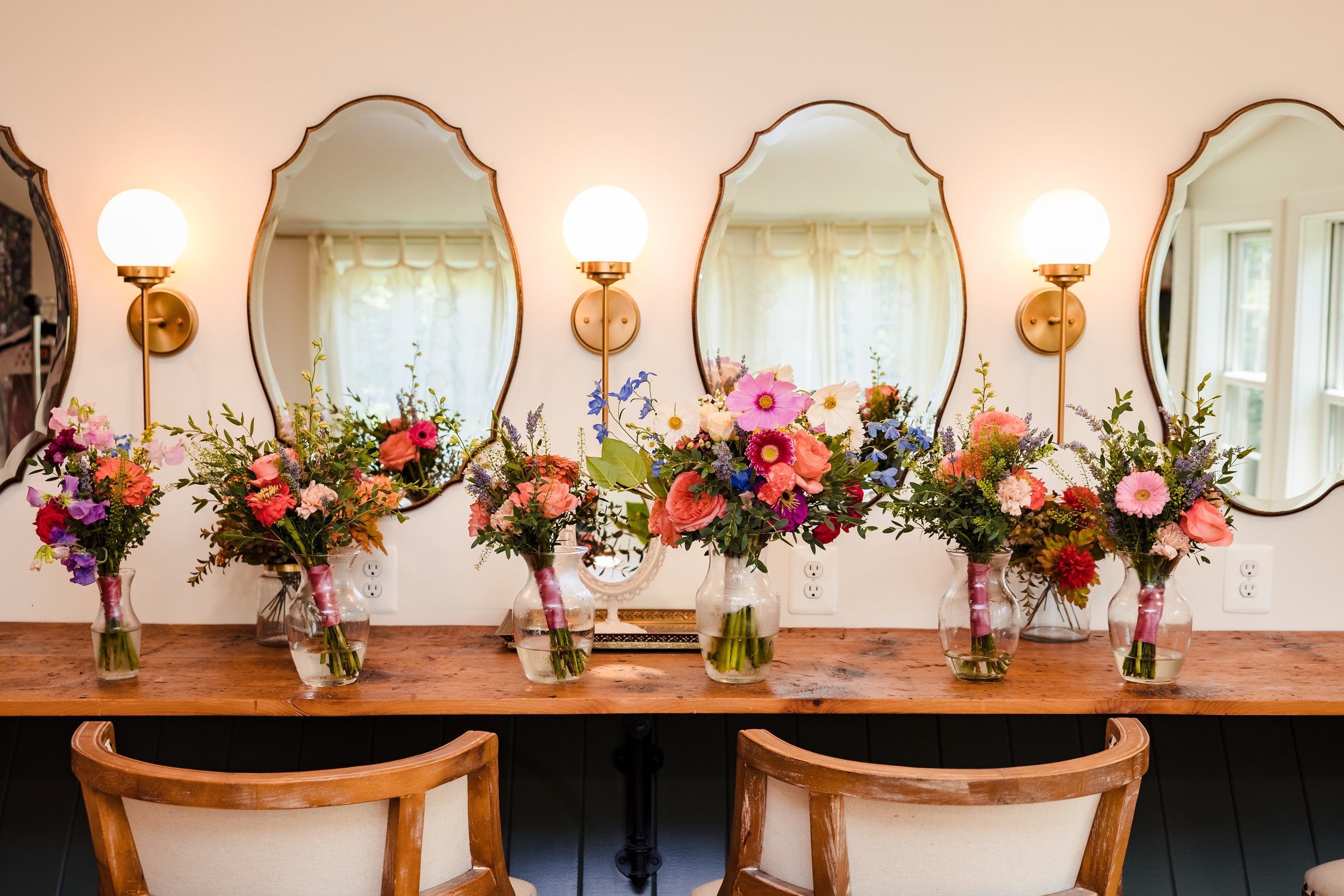 A wooden table decorated with eight glass vases filled with colorful flowers including pink, purple, and white blossoms. Behind the table, there are four large, ornate mirrors with curved edges mounted on a white wall. The mirrors reflect a bright ro