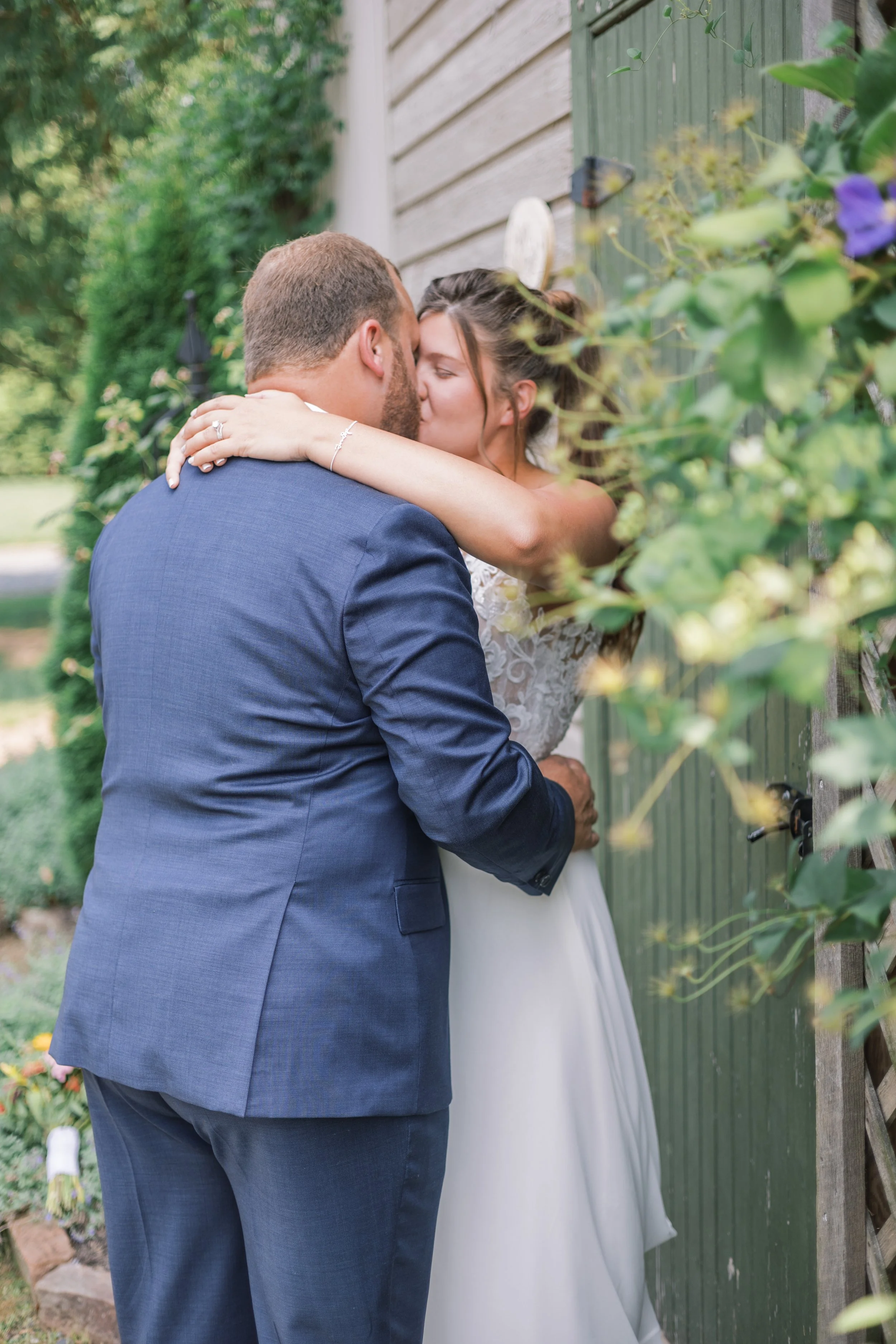 A couple shares a kiss outdoors, with the woman in a white dress and the man in a blue suit, near a green wooden fence and lush greenery.