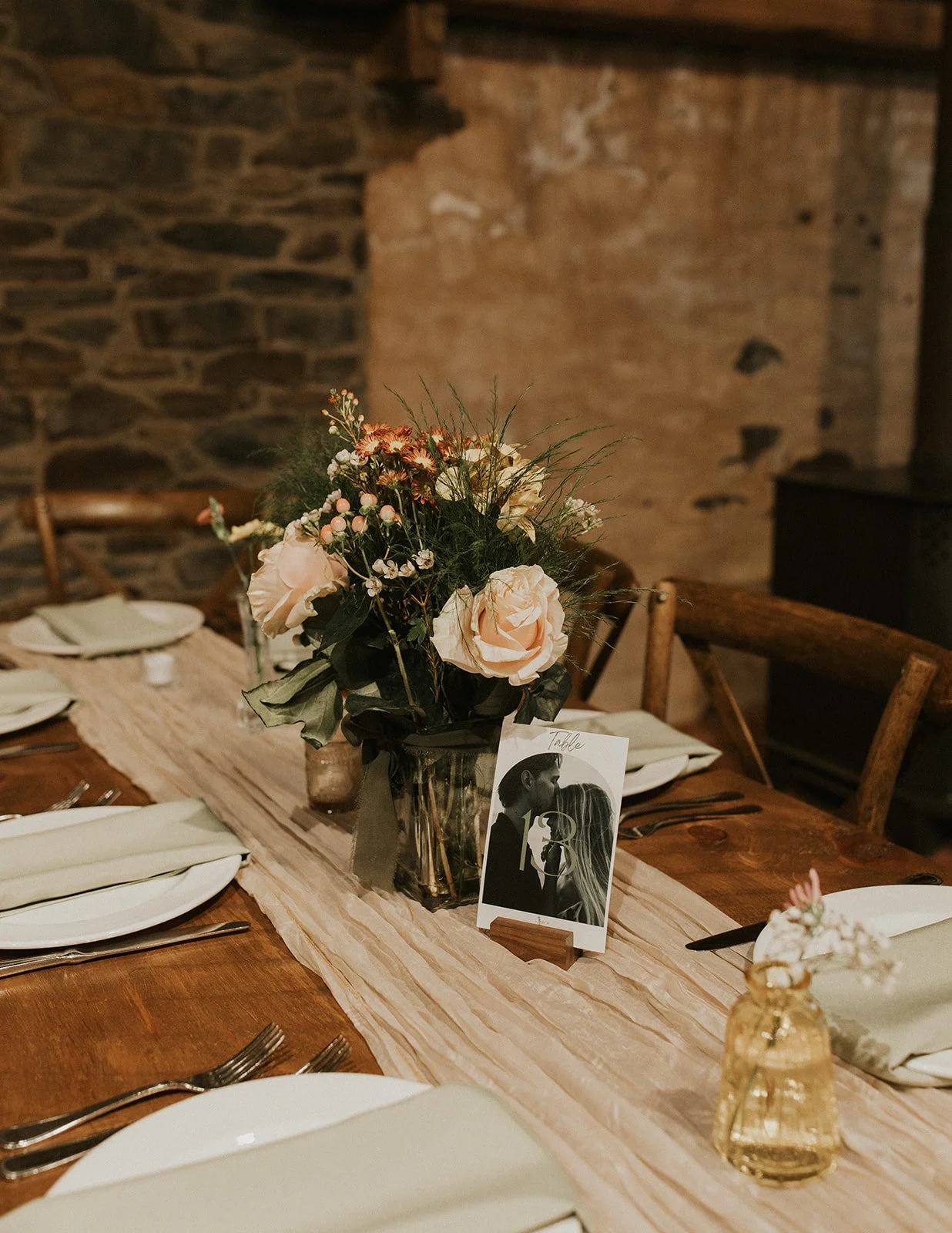A wooden dining table set for a wedding or special event, decorated with a beige table runner, white plates, and silverware. In the center, there is a floral arrangement with large white roses, small pink flowers, and greenery in a black vase, along 