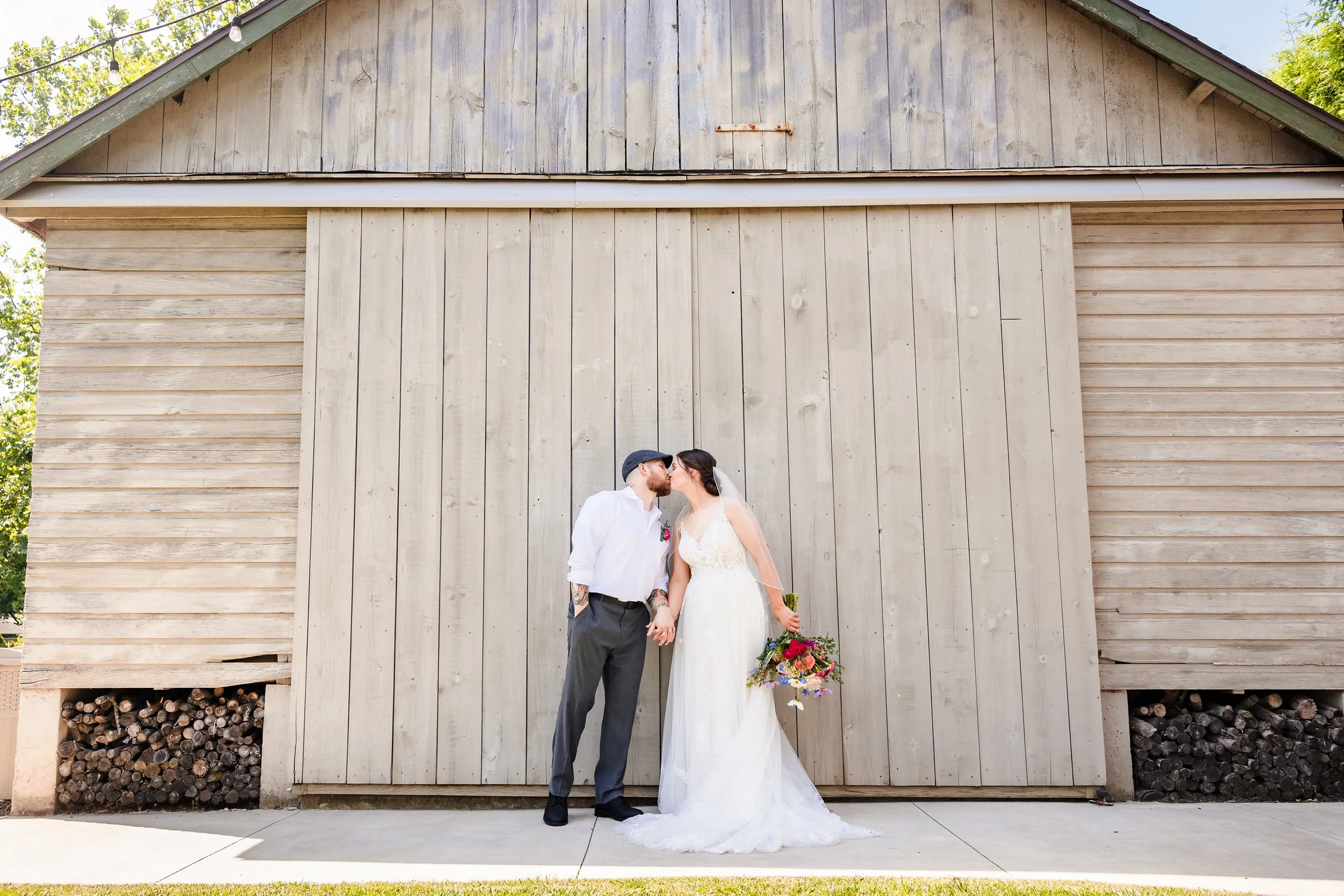 A bride and groom are standing in front of a rustic wooden barn, kissing. The bride is wearing a white wedding dress and holding a colorful bouquet, while the groom is dressed in a gray suit, wearing a cap, and has tattoos on his arm.