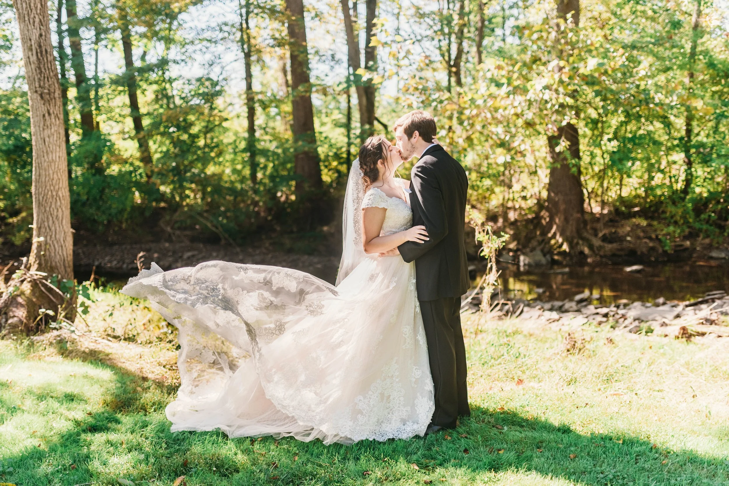 A bride and groom share a kiss outdoors in a wooded area during their wedding, with sunlight filtering through the trees.
