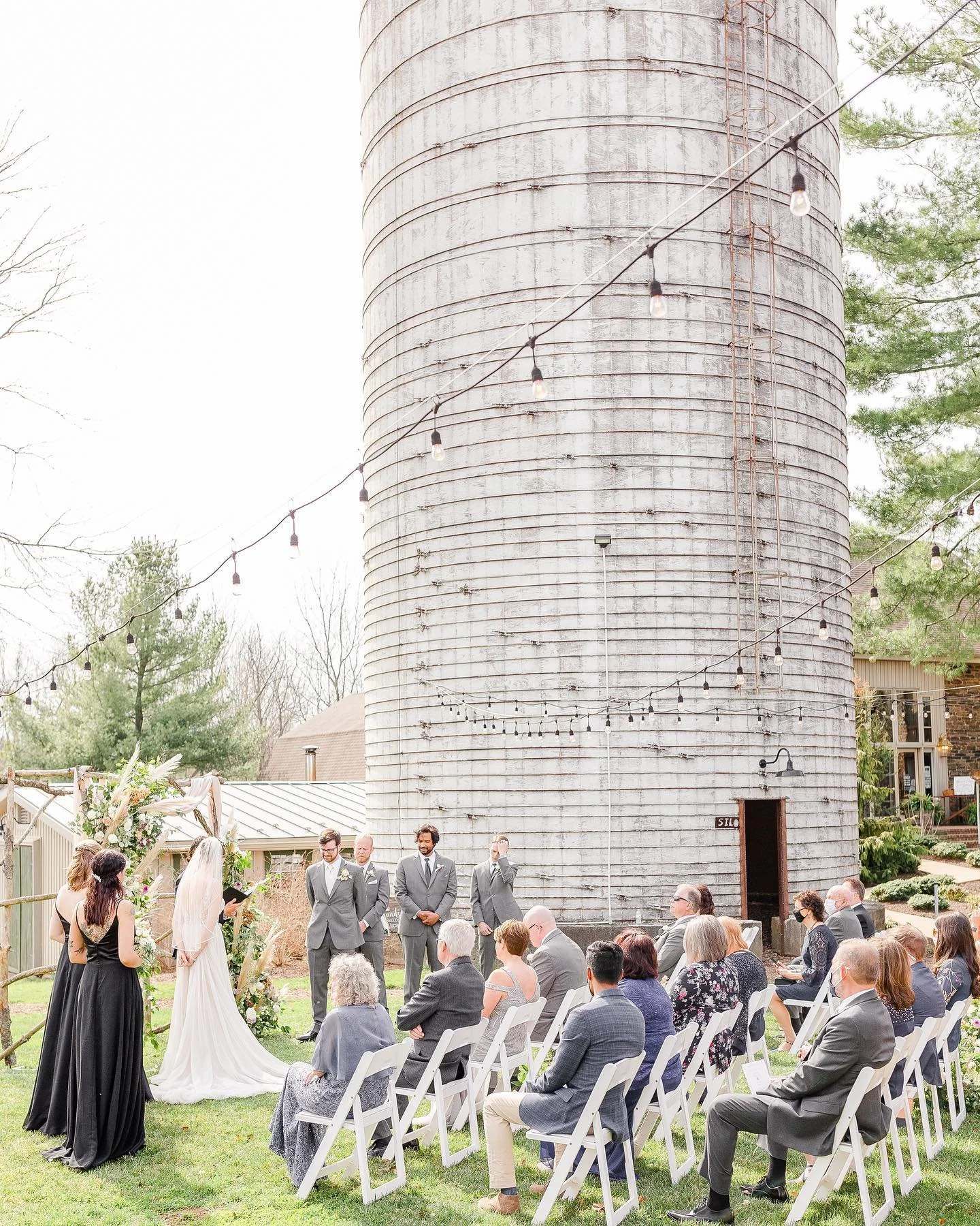 Outdoor wedding ceremony taking place in front of a tall, weathered barn silo with string lights overhead. The bride and groom stand at the altar with officiant, surrounded by guests seated in white chairs. The bride is wearing a white gown with a ve