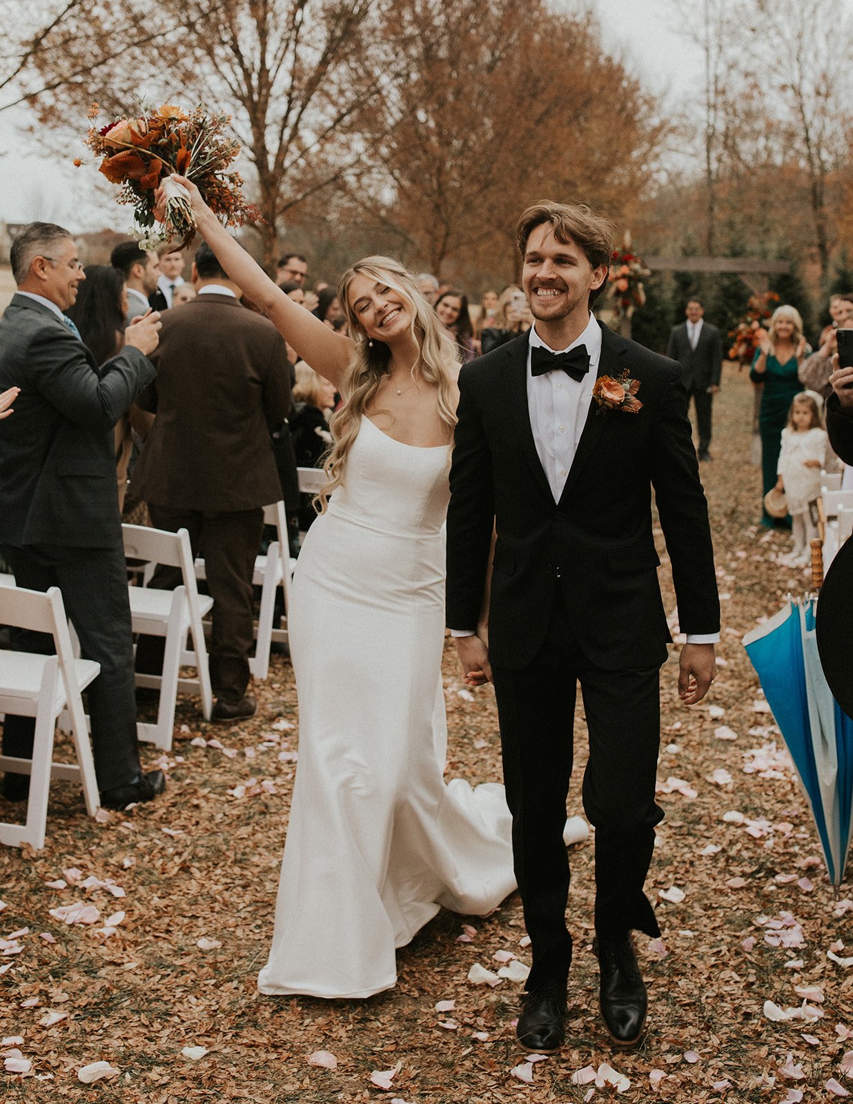 A bride and groom walking hand-in-hand at their outdoor wedding ceremony with guests and fall foliage in the background.