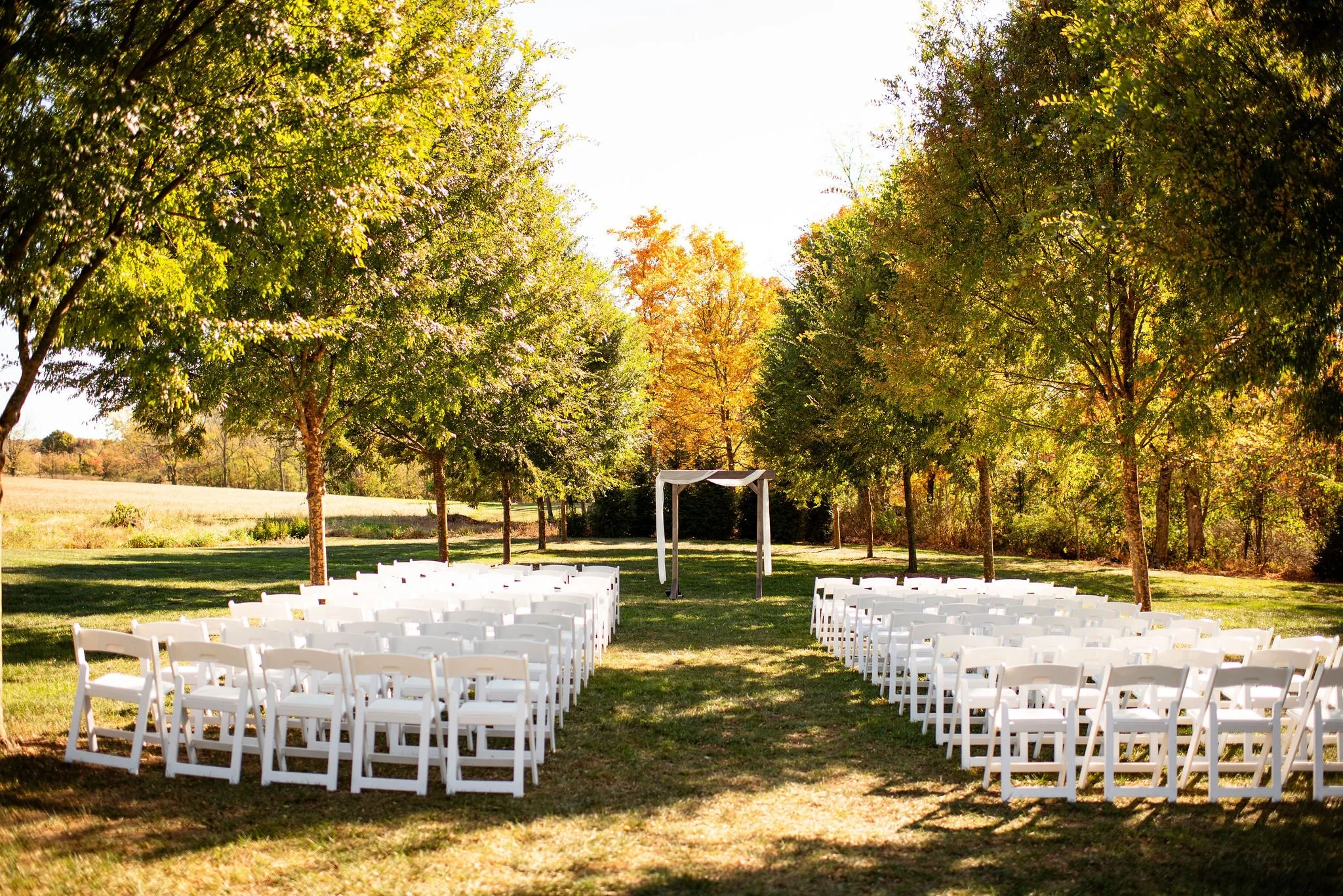Outdoor wedding ceremony setup with white chairs arranged in rows on grass, a white arch for the officiant, surrounded by trees with fall foliage.