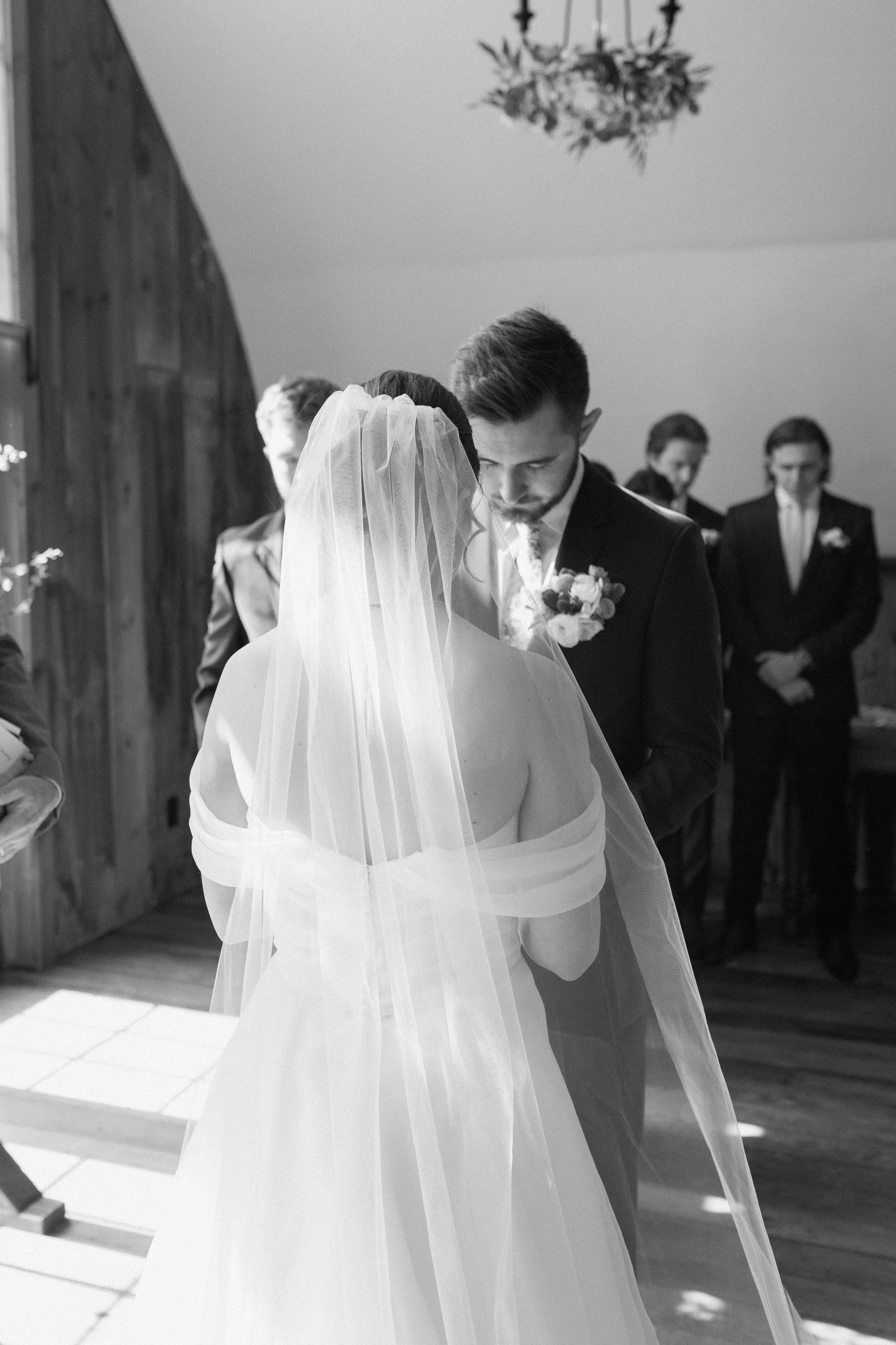 A bride and groom standing during a wedding ceremony indoors, with bridesmaids and groomsmen in the background. The bride is wearing a veil, and the groom is dressed in a suit holding a bouquet of flowers.
